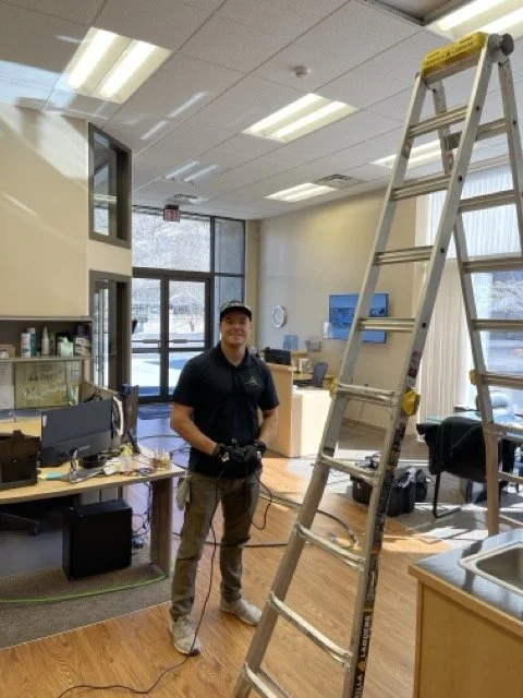 Man standing in an office with a ladder, computer desk, and window.