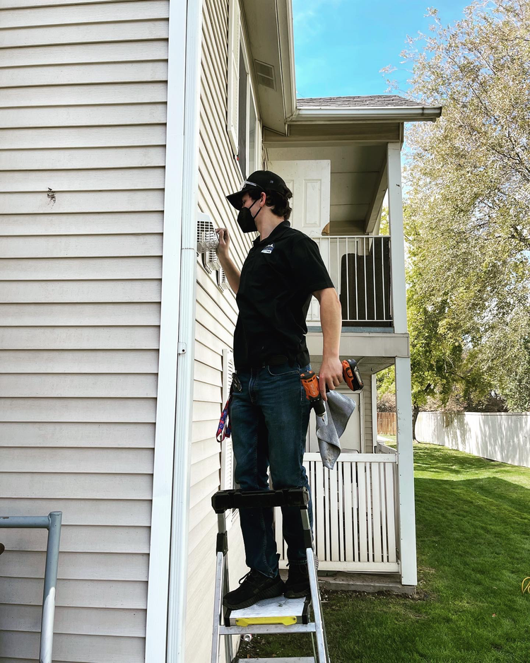 A technician standing on a step ladder working on a house's exterior, wearing a black mask, black shirt, jeans, and a cap, with tools attached to her belt and holding a cloth.