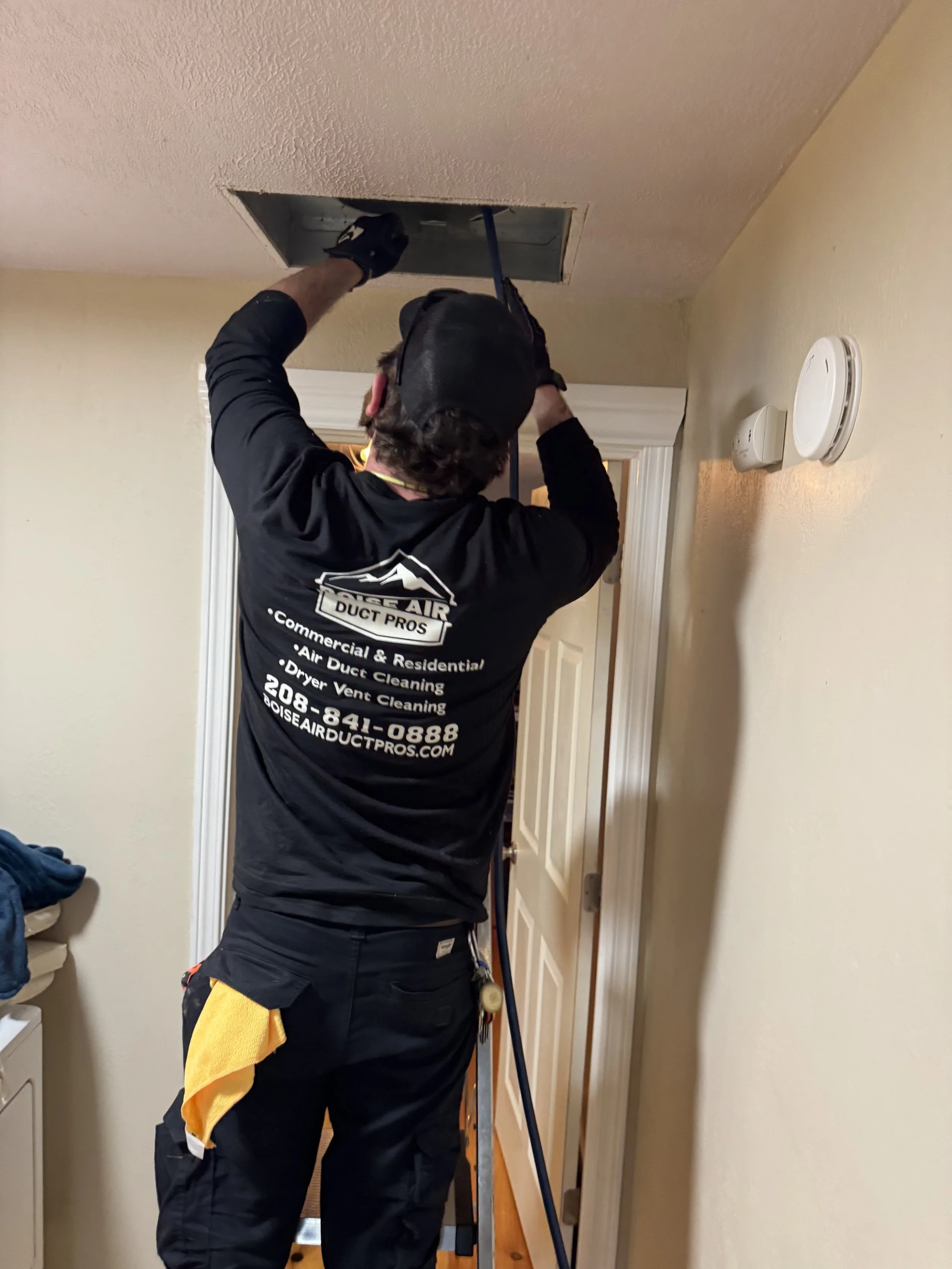 A technician is repairing or inspecting an attic access hatch in a ceiling, standing on a ladder, wearing a black uniform with company information on the back.