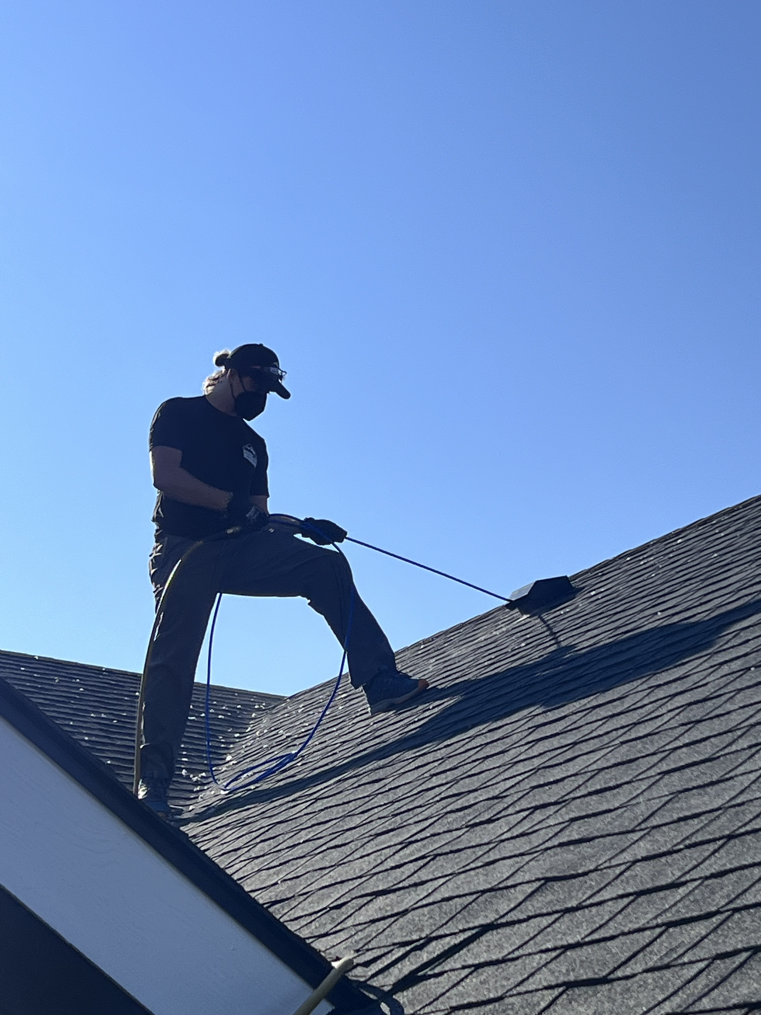 A person wearing a black cap, black shirt, and gray pants cleaning a sloped roof with a power washer under clear blue sky.