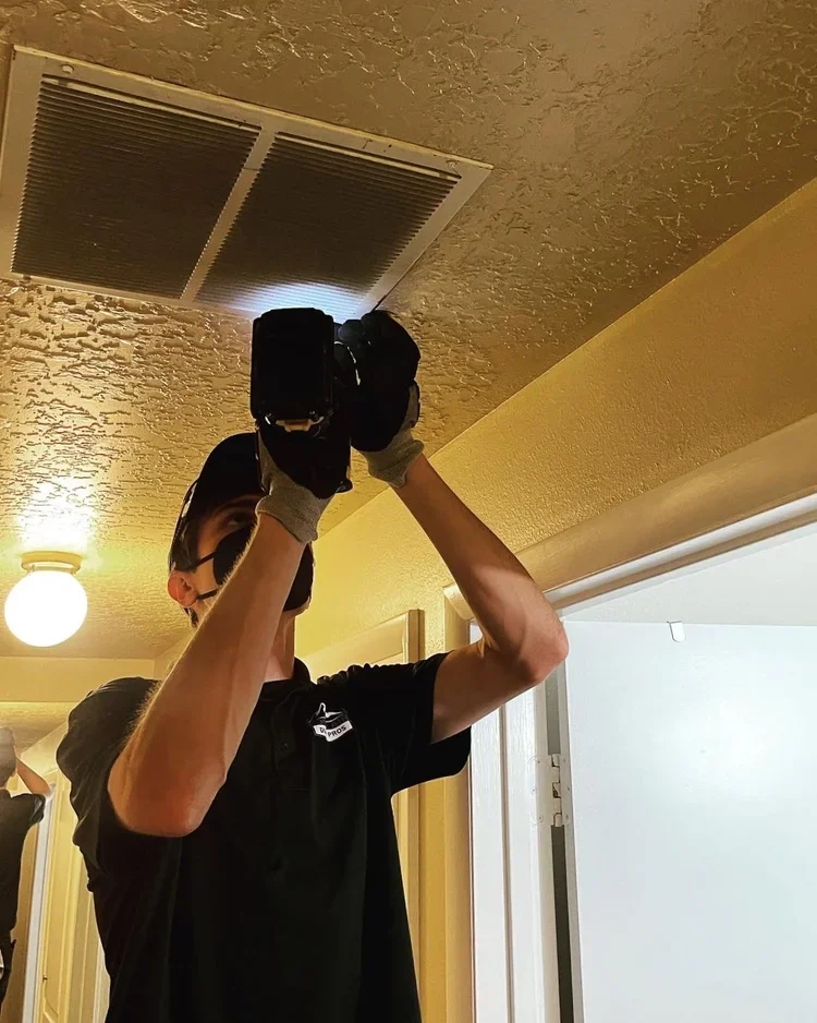 A person wearing gloves, a cap, and a black shirt is taking a photo of a ceiling vent with a camera or phone inside an indoor room.