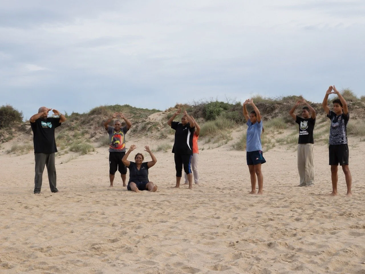 A group of people practicing yoga on a sandy beach, with some standing and others sitting or kneeling, under cloudy skies.