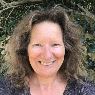 A woman with curly hair smiling outdoors with green foliage in the background.
