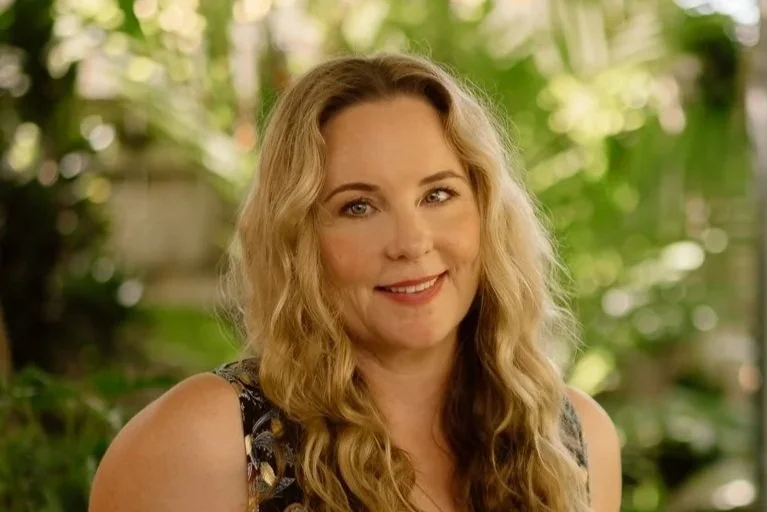 A woman with long, wavy blonde hair smiling at the camera, outdoors with blurred green foliage in the background.