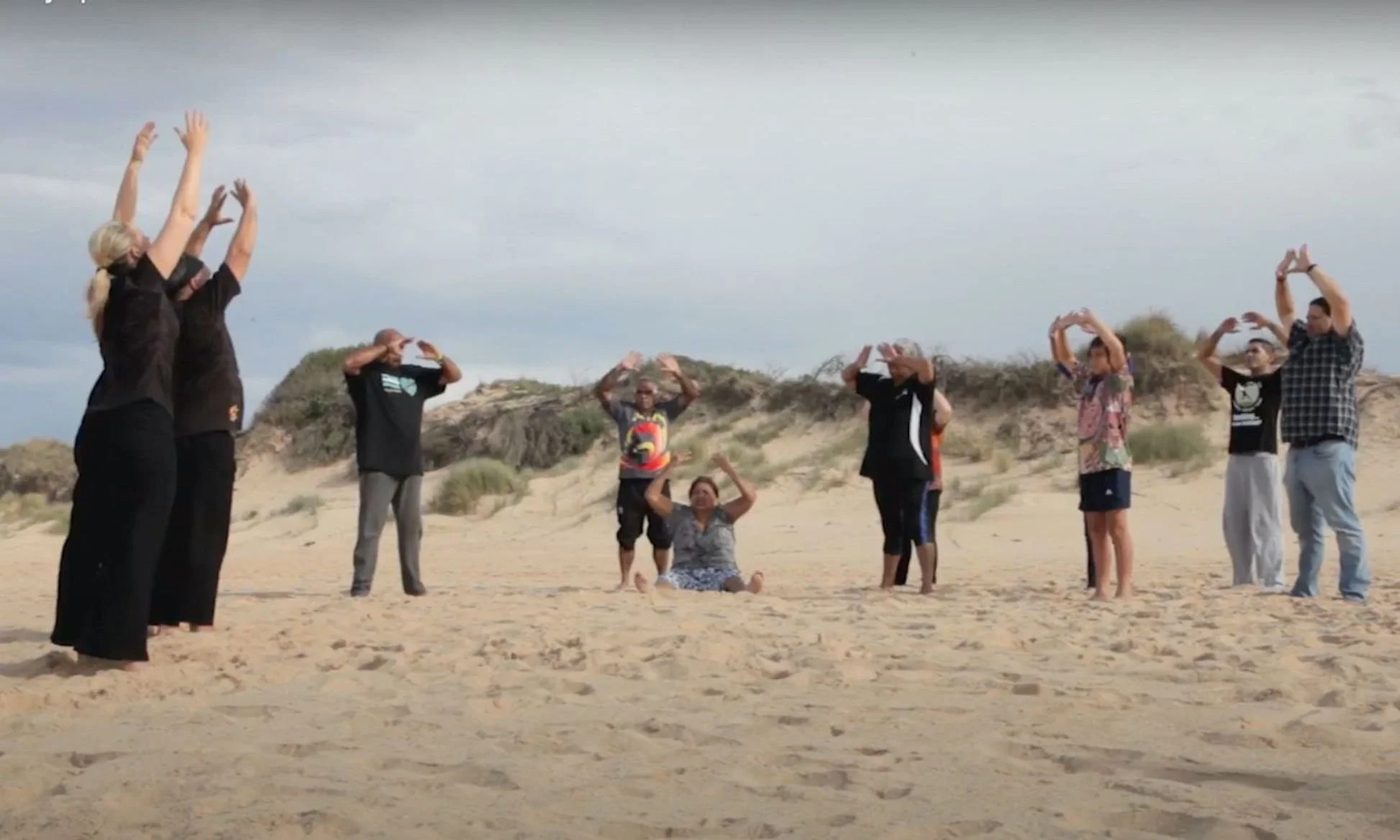 A group of people practicing yoga on a sandy beach, with some standing and others sitting or kneeling, under cloudy skies.