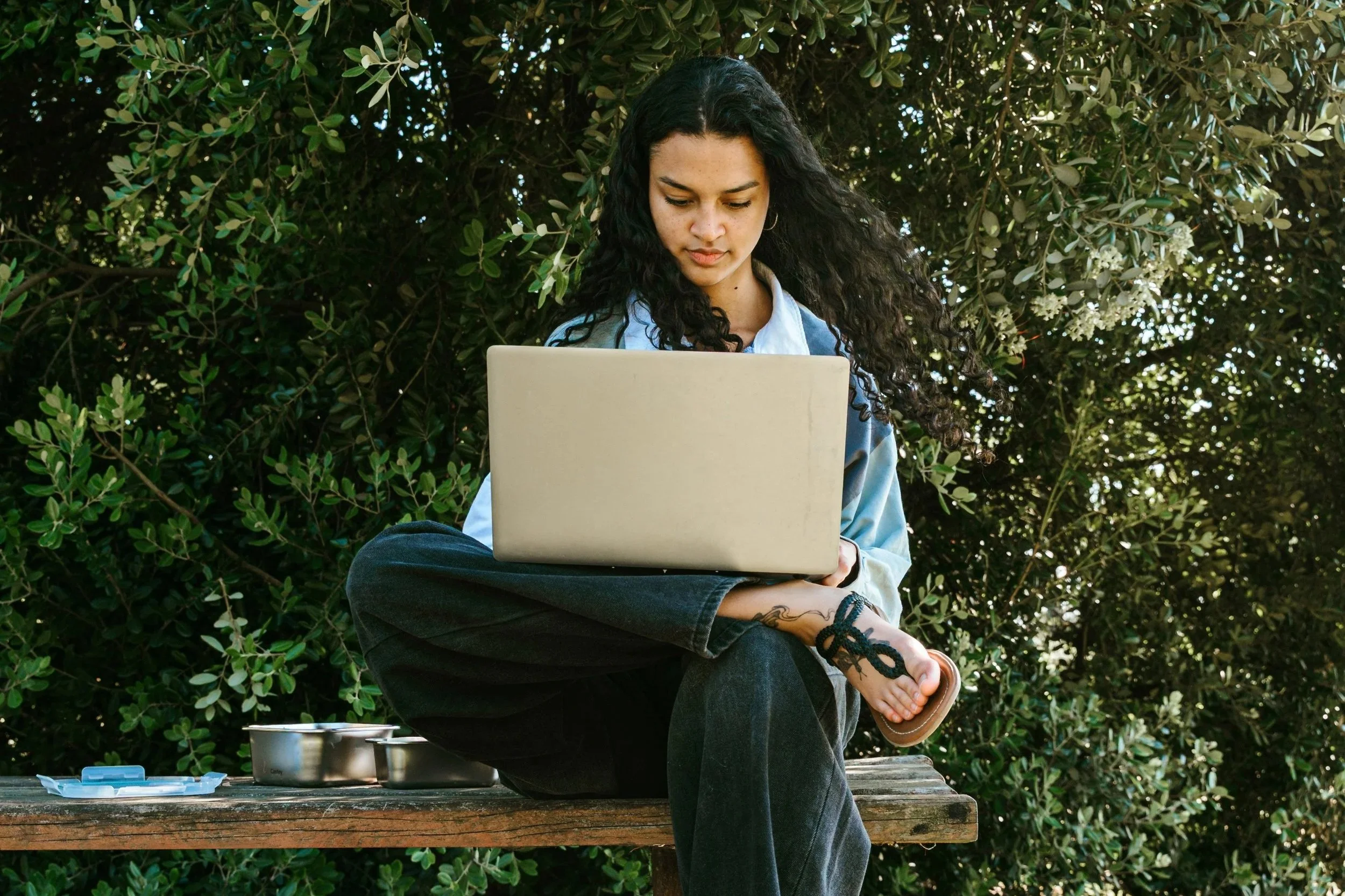 A young woman with long, curly dark hair sitting outdoors on a wooden bench, working on a laptop surrounded by greenery.