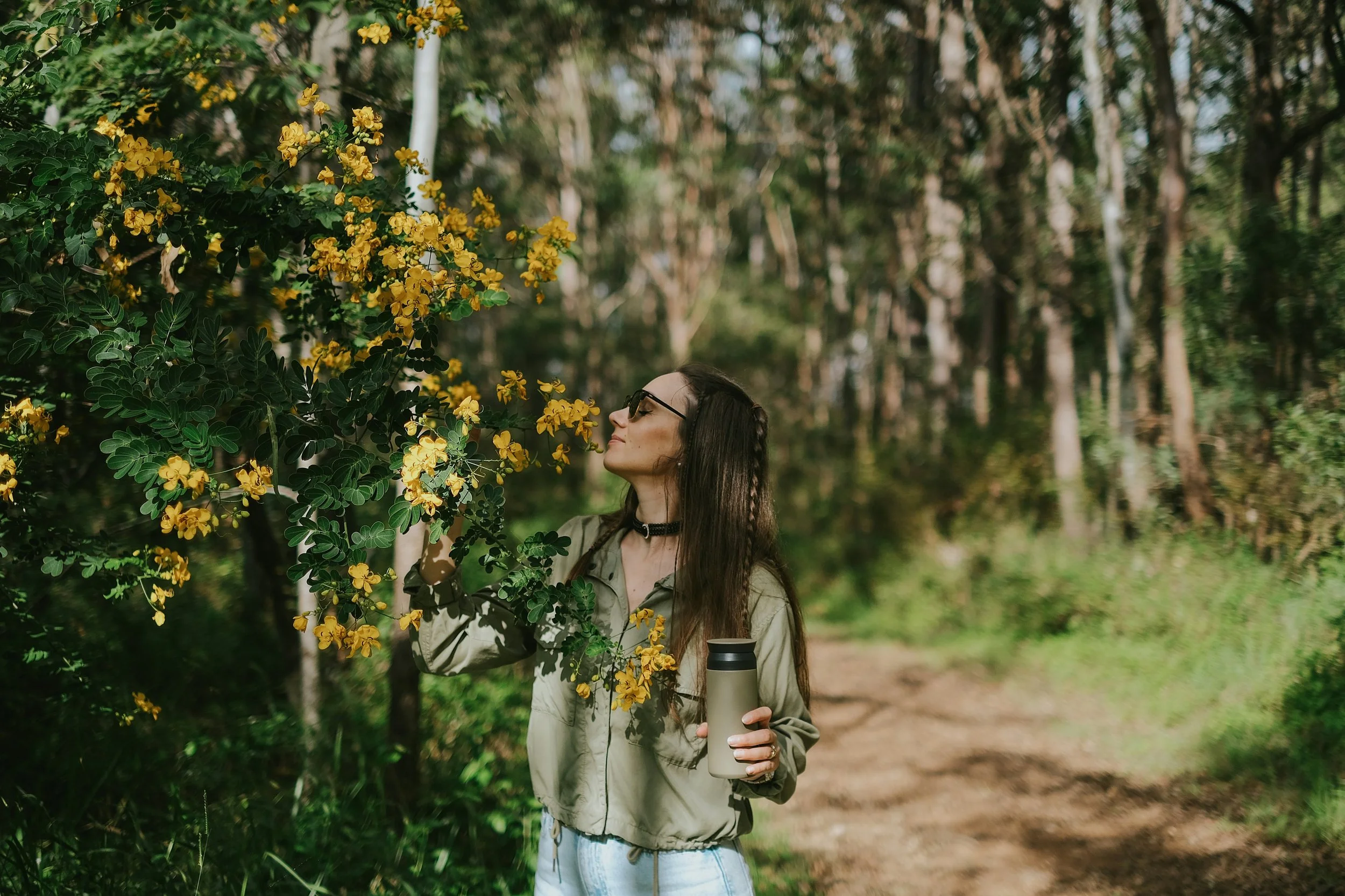 A woman with long brown hair, wearing sunglasses, a beige jacket, and light blue jeans, stands in a forested area holding a plant with yellow flowers and a thermos.