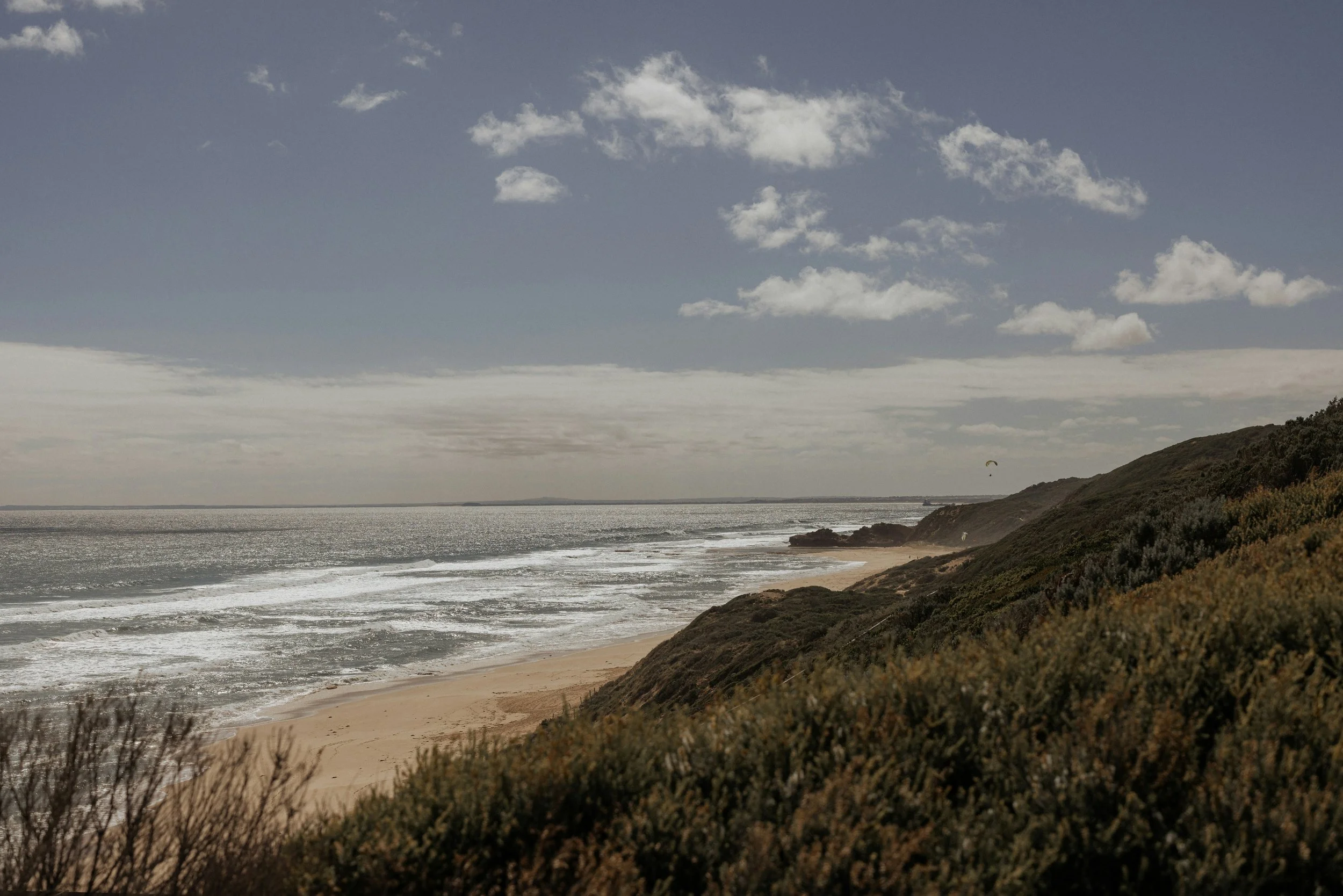 Coastal landscape with sandy beach, ocean waves, green hillside, and a scattered cloudy sky.