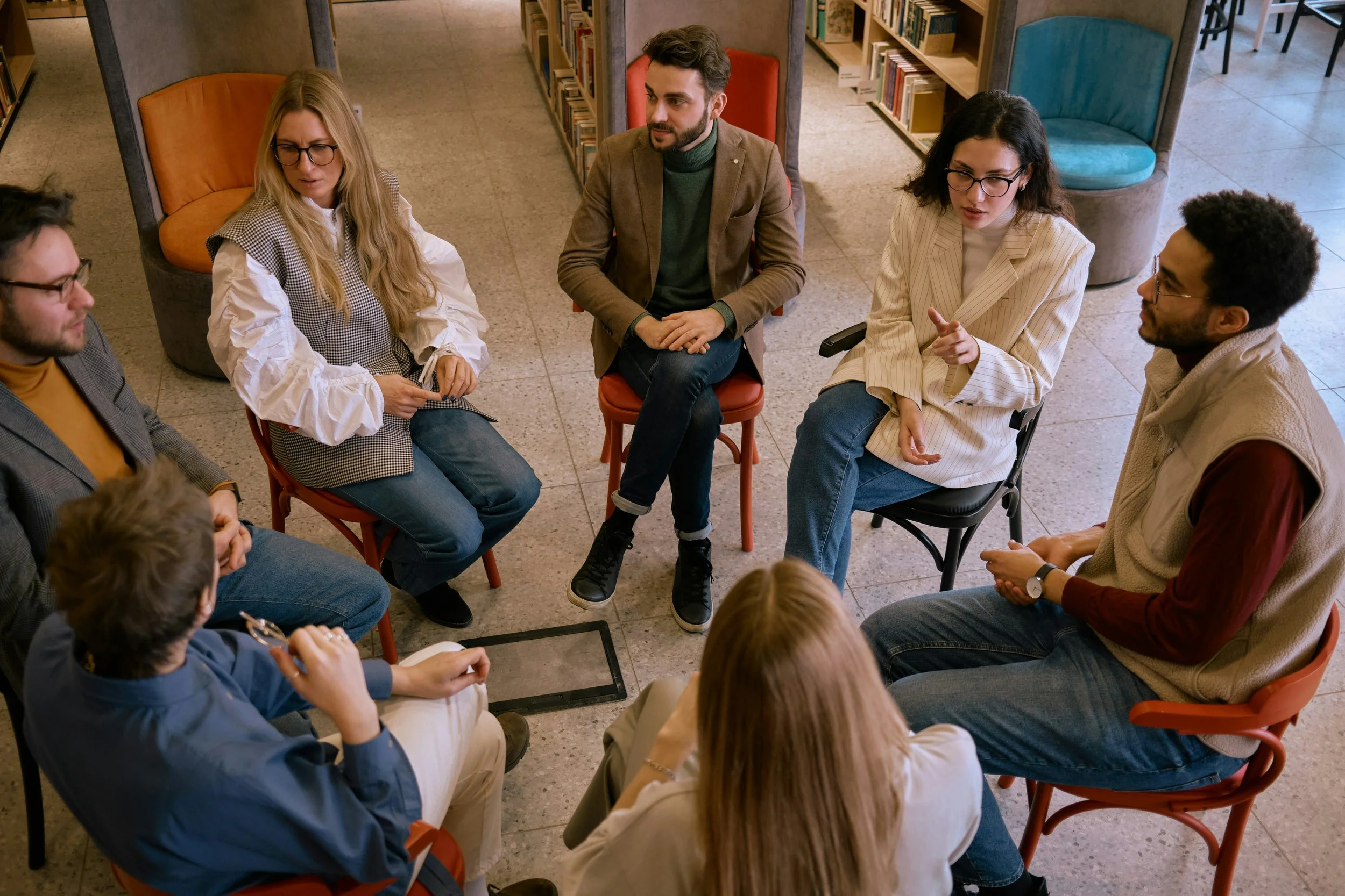A group of six people sitting in a circle in a library, engaged in a discussion. The room has bookshelves and cozy chairs.