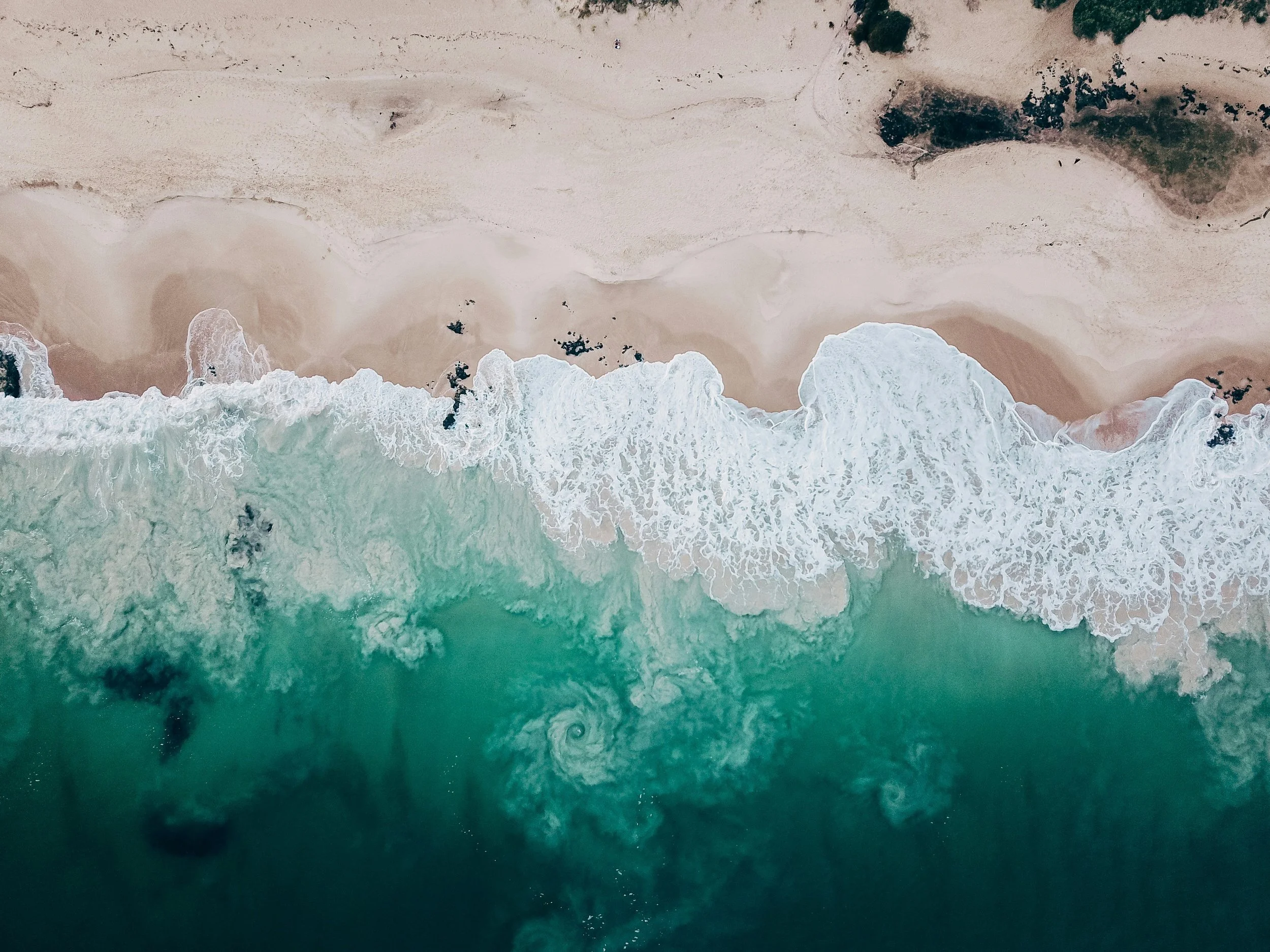 An aerial view of sandy beach and ocean waves crashing onto the shore with dark rocks and vegetation at the top of the image.