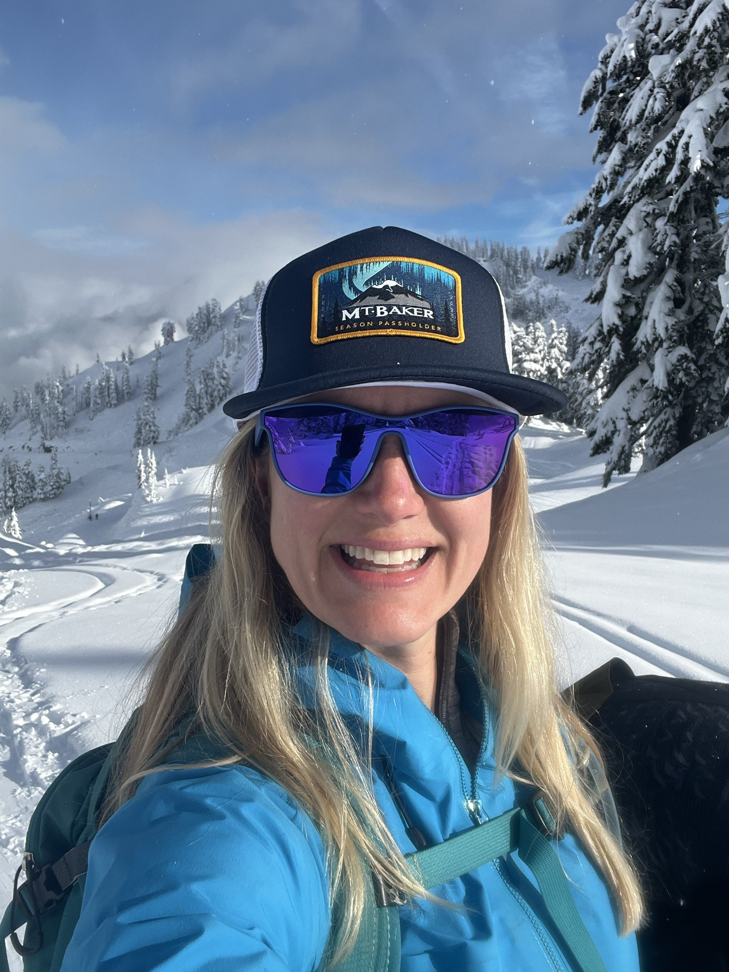 A woman smiling outdoors in a snowy mountain landscape, wearing a blue jacket, purple sunglasses, a black cap with a Mountain logo, and a backpack.