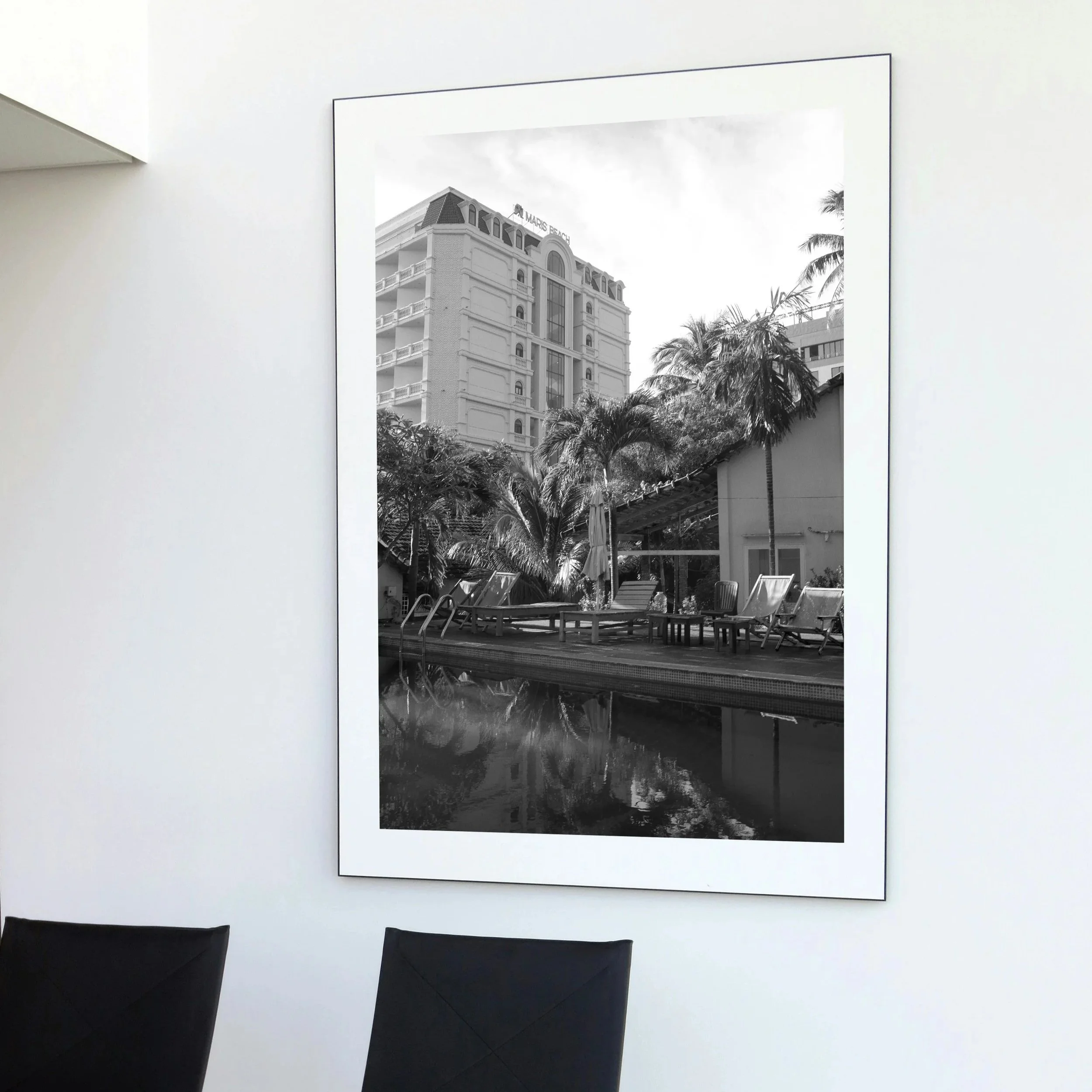 Black and white photograph of a hotel building with palm trees, a pool area with lounge chairs, and reflections in the water. Wall Print