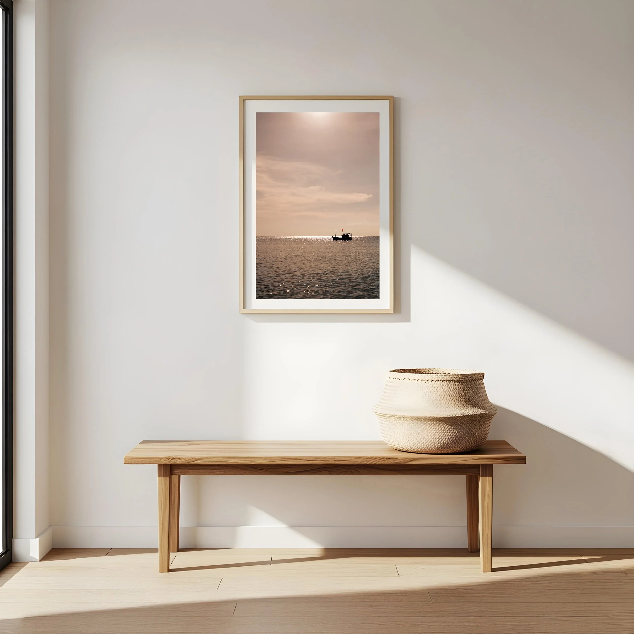 Minimalist interior with a light wood bench, a woven basket, white wall, framed seascape photo of a boat on the water at sunset, and sunlight casting shadows. Island Wall print