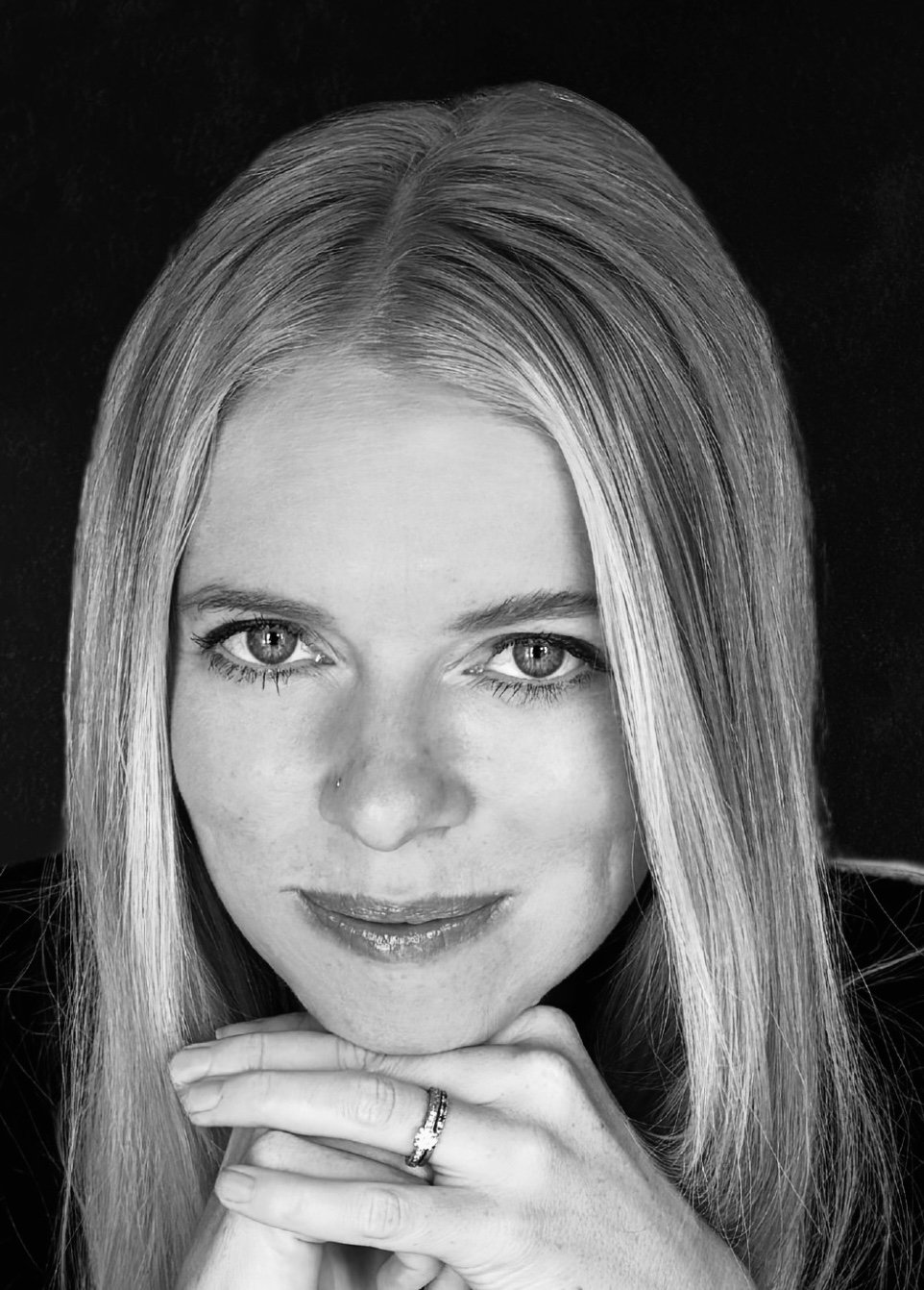 Black and white portrait of a woman with long straight hair,-looking directly at the camera, resting her chin on her hands, showing rings on her fingers.