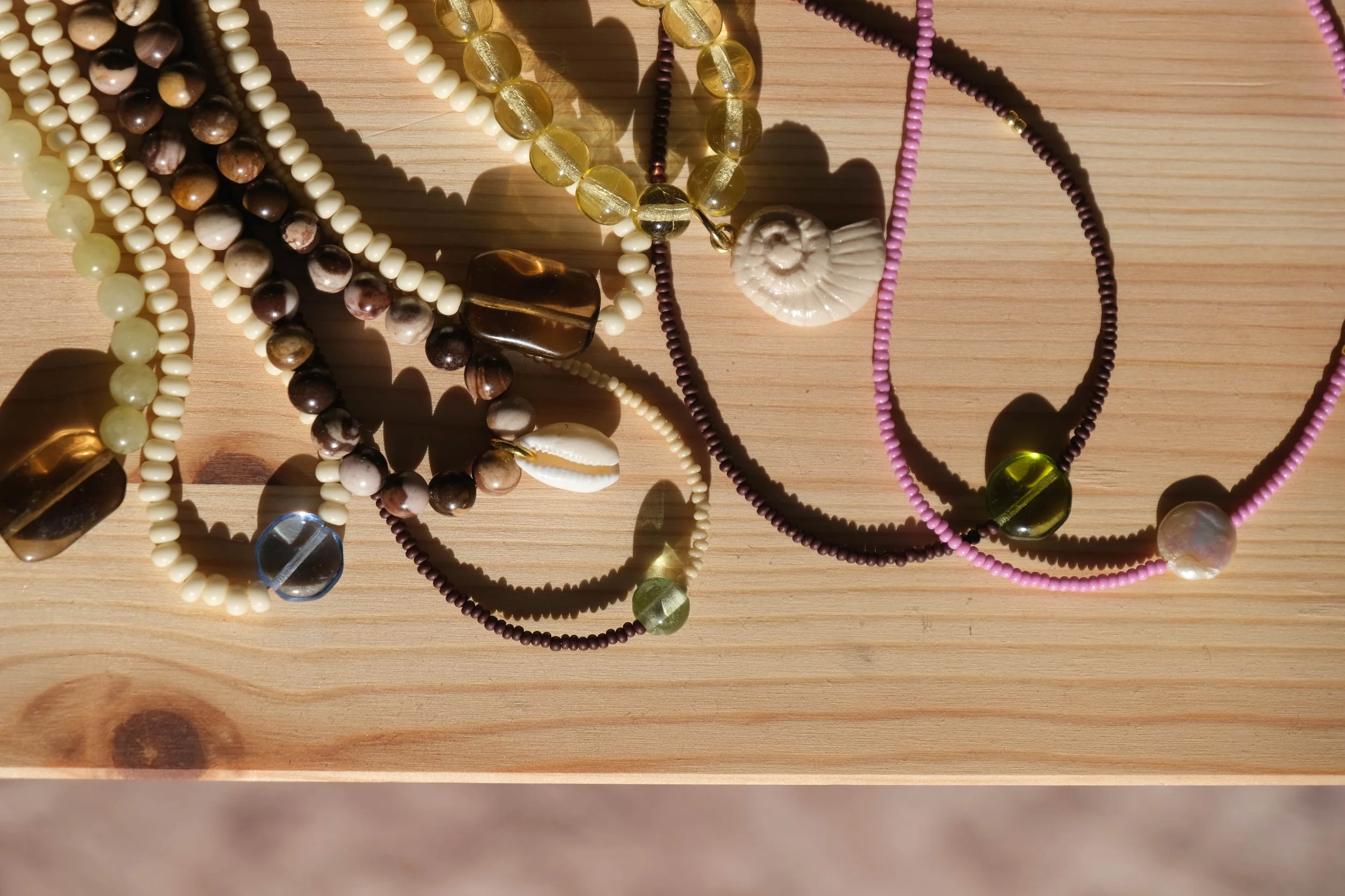 A close-up of various beaded necklaces and bracelets laid on a wooden surface, featuring different colors and sizes of beads including white, brown, yellow, pink, purple, and green.