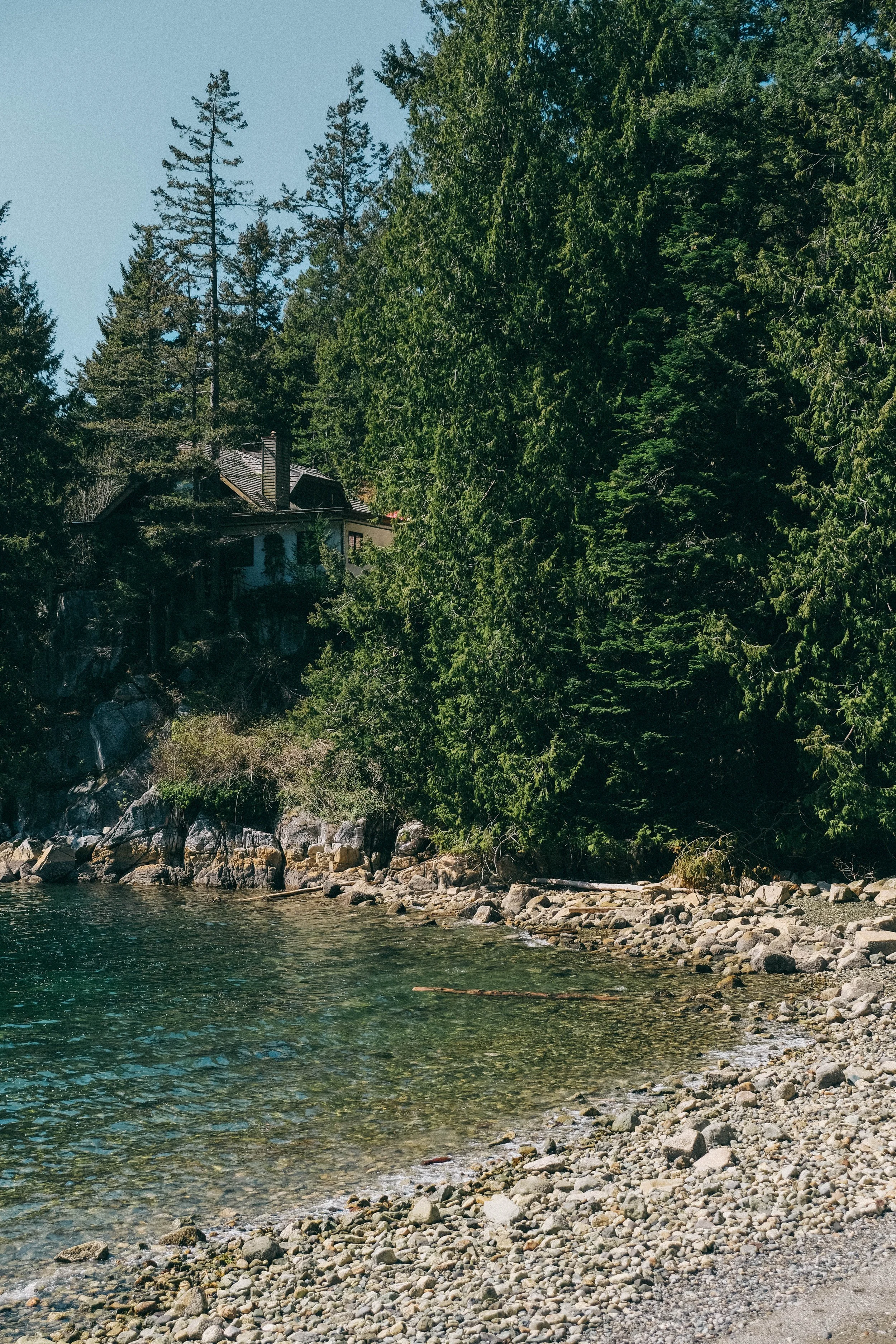 A lakeside scene with clear water, a rocky shoreline, and dense green trees. Partially visible house on the hillside among trees and a rocky cliff.