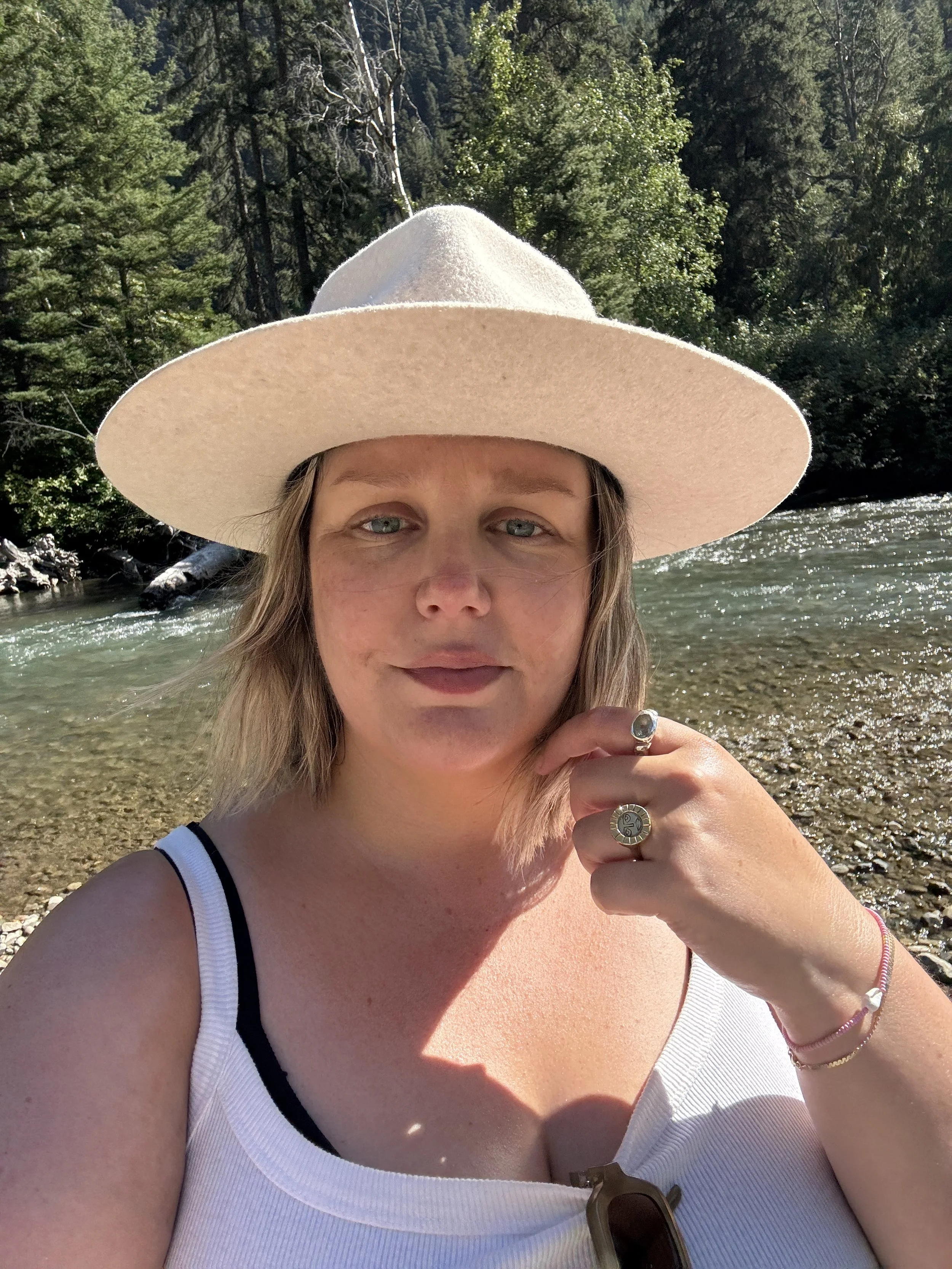 A woman wearing a wide-brimmed hat, a white tank top, and jewelry, standing in front of a river with trees and mountains in the background, on a sunny day.