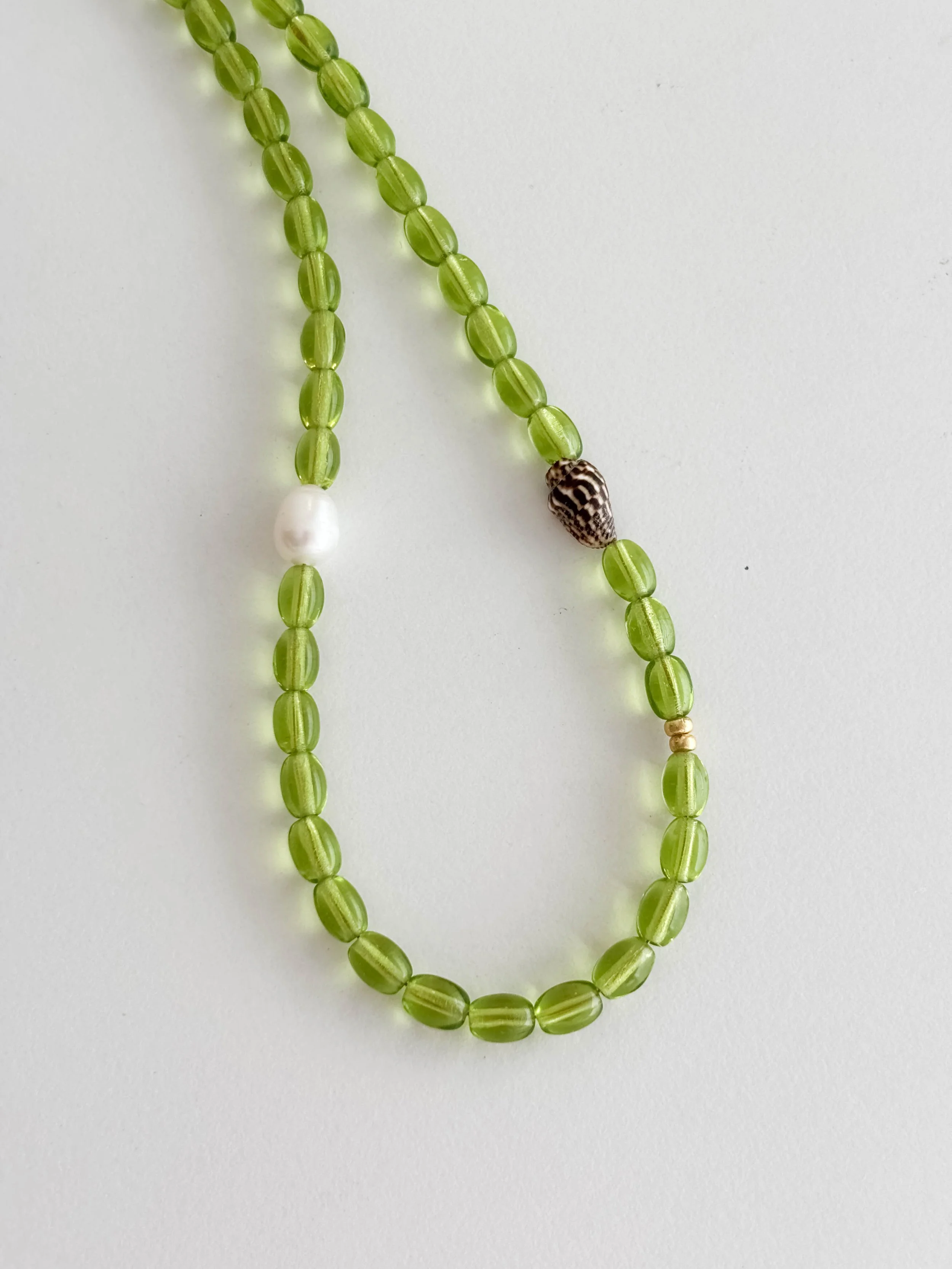 Green beaded necklace with a single white pearl and a small black and white shell clasp placed on a white background.