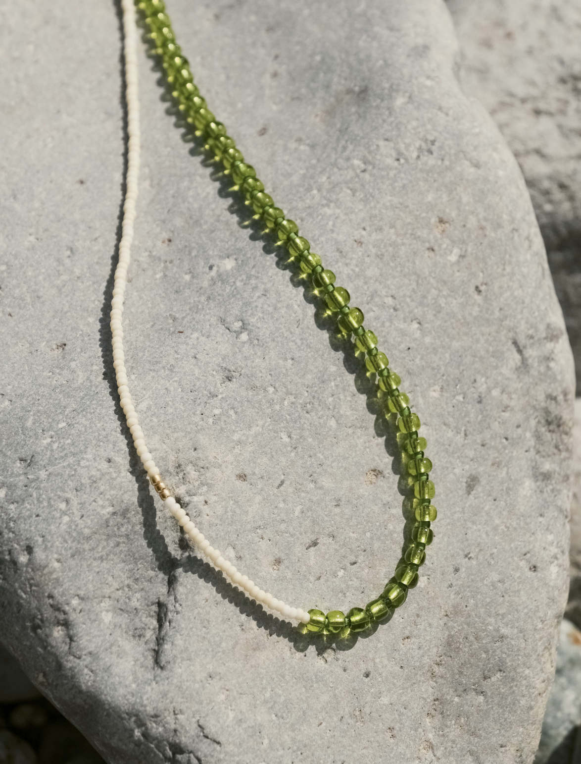 Two beaded necklaces, one white and one green, lying on a gray stone surface outdoors.
