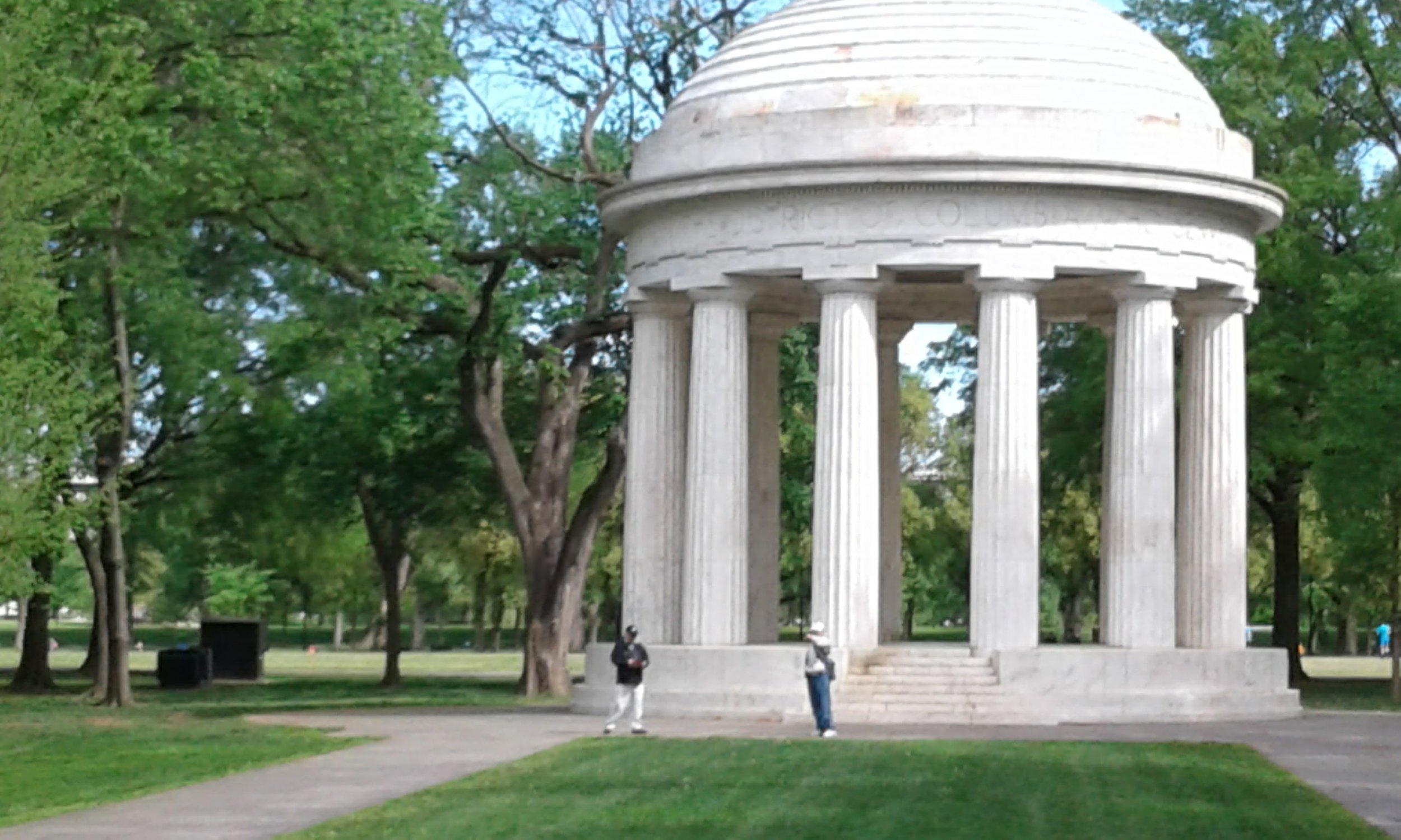 A white marble dome shaped monument with classical columns in a park with green grass and trees, and three people standing near it.