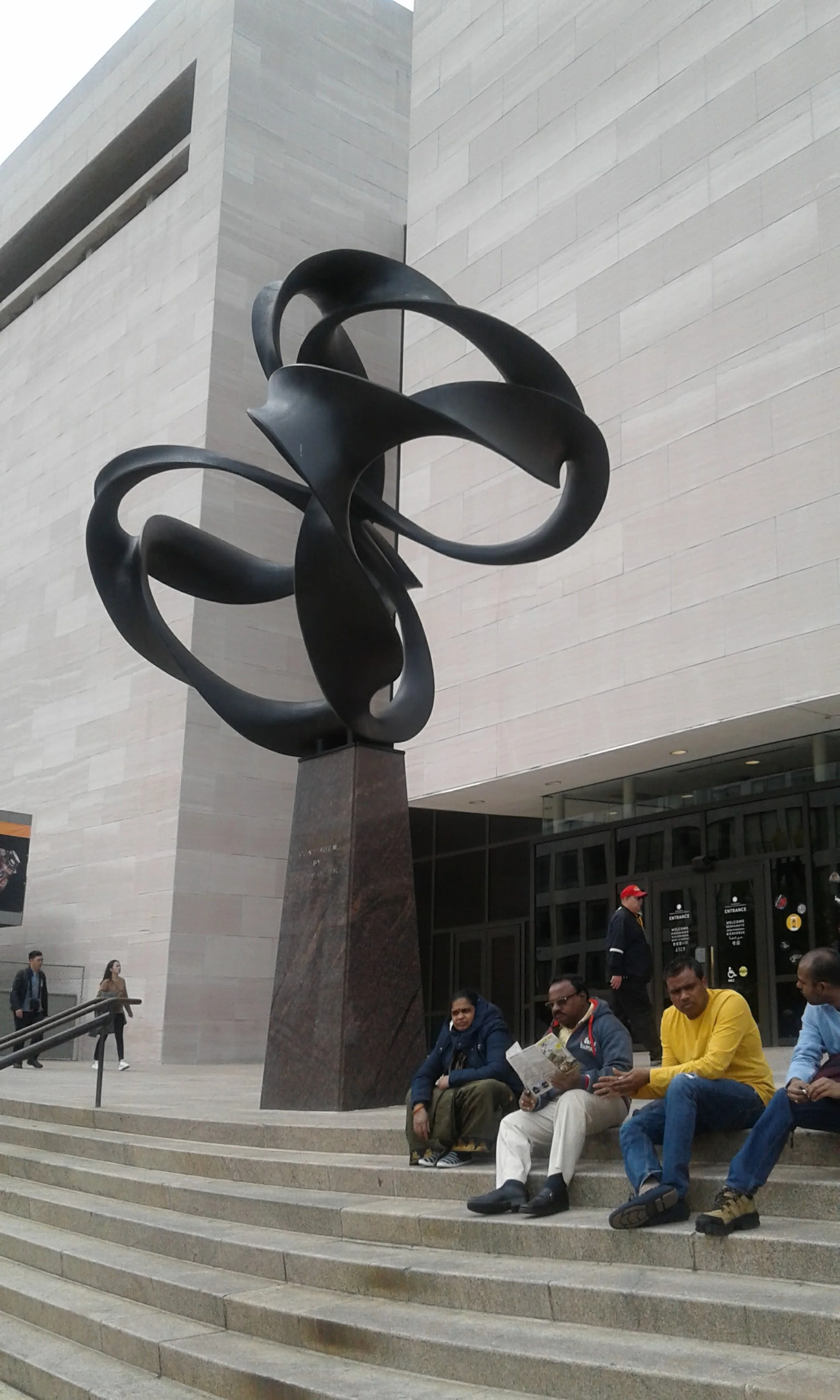 An outdoor sculpture on a tall dark base with twisting, looping black metal rods, in front of a modern building with stone exterior and glass entrance doors. Several people are sitting on the steps in front of the sculpture, and others are walking ne