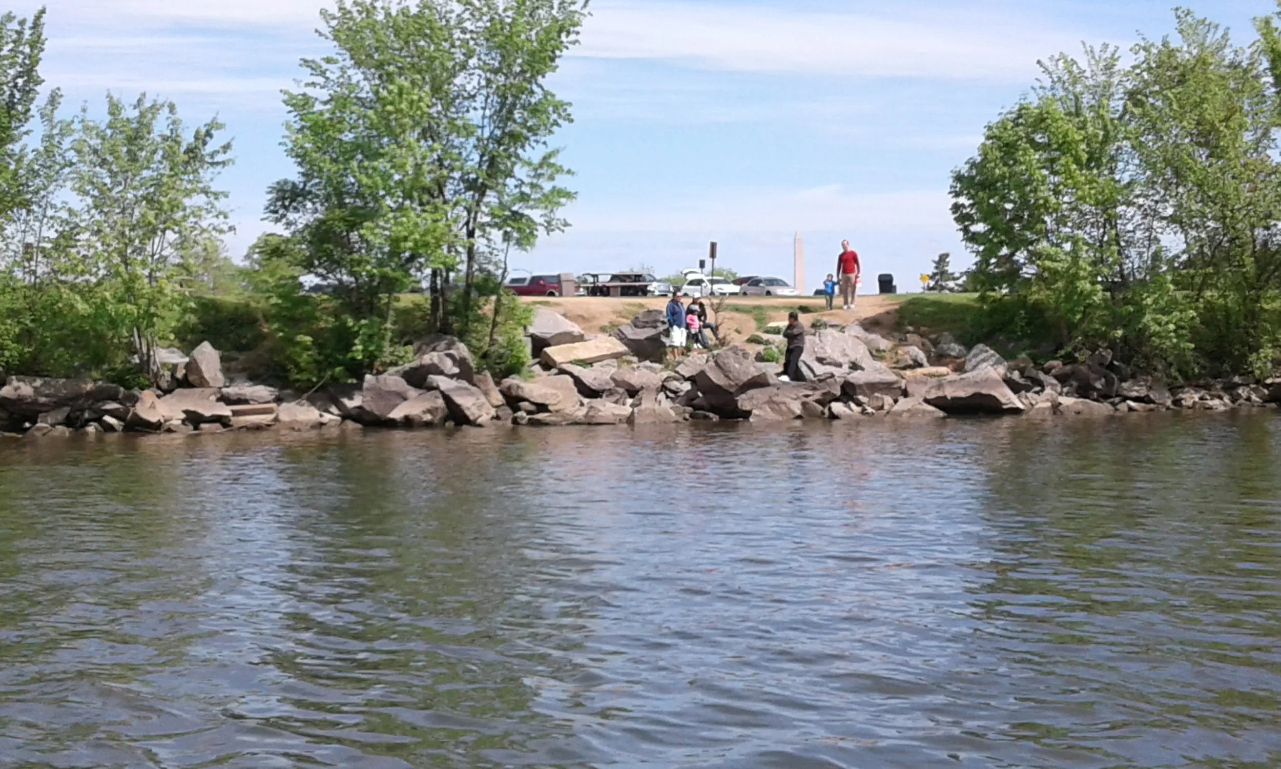 View of a riverbank with people exploring rocks near the water, trees on the sides, and parked cars and a monument in the background.