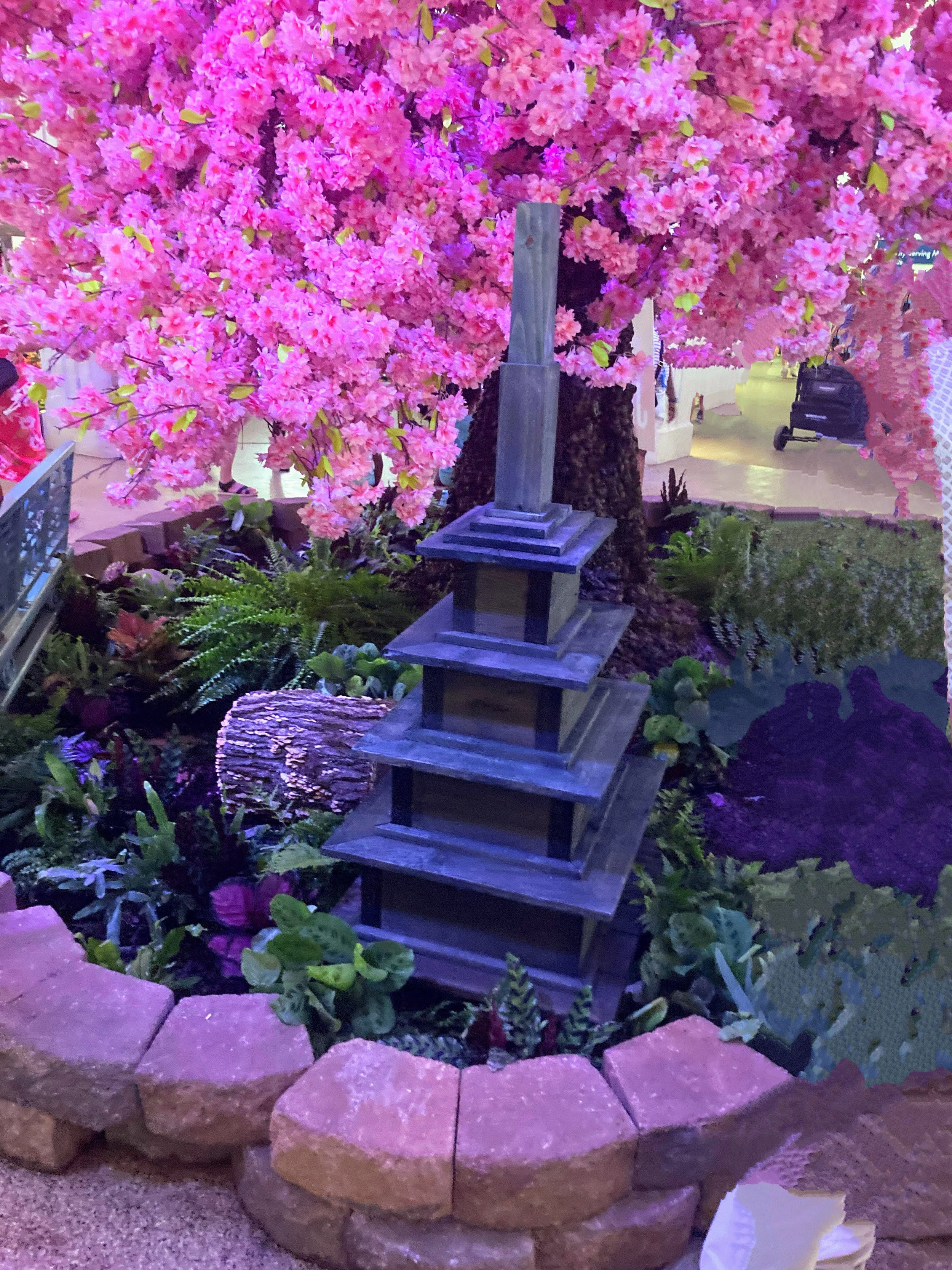 Artificial cherry blossom tree with pink flowers, surrounding a wooden pagoda-style structure, in an indoor setting with plants and decorative stones.