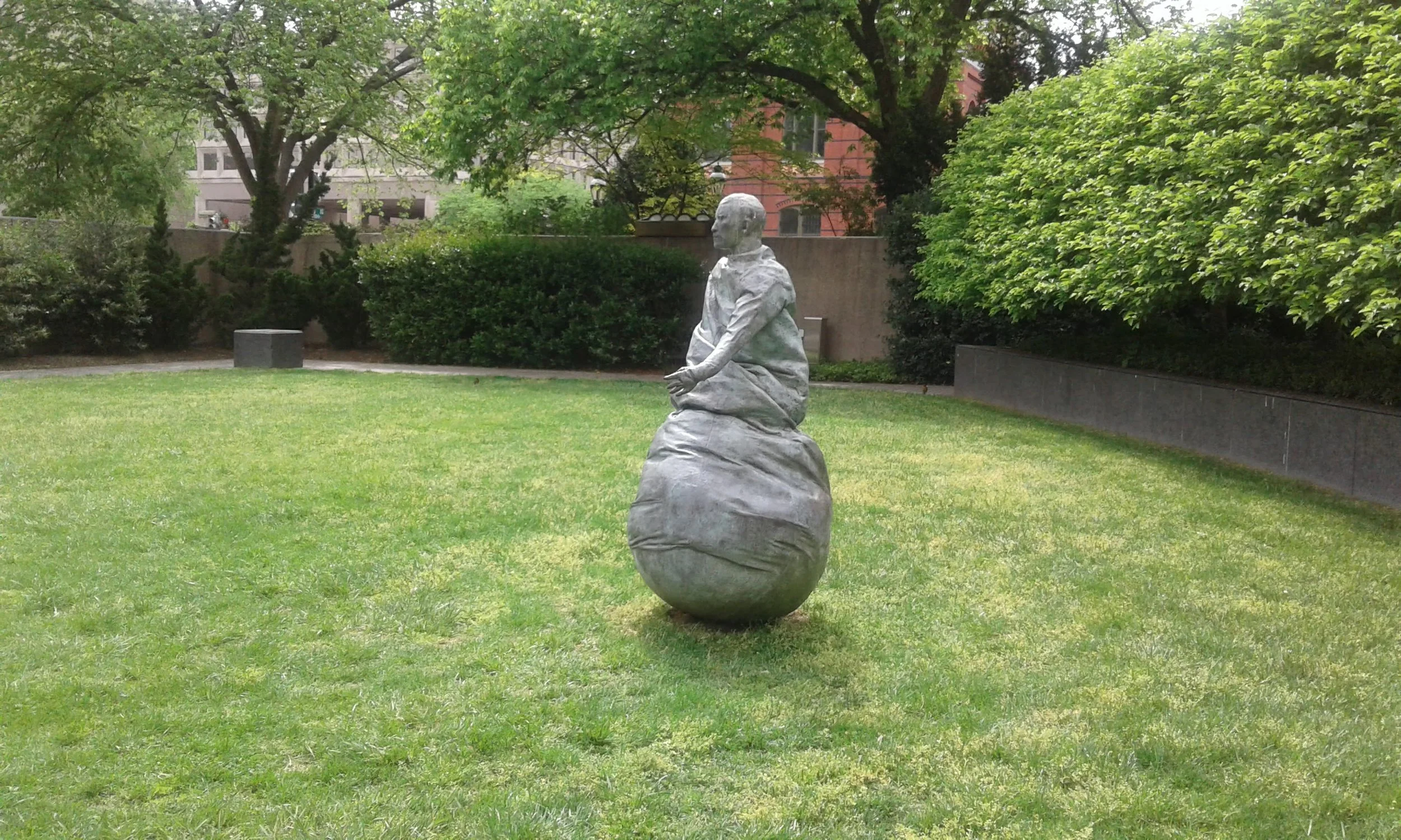 A sculpture of a seated person on top of a rounded base, placed in a grassy park with trees and bushes in the background.