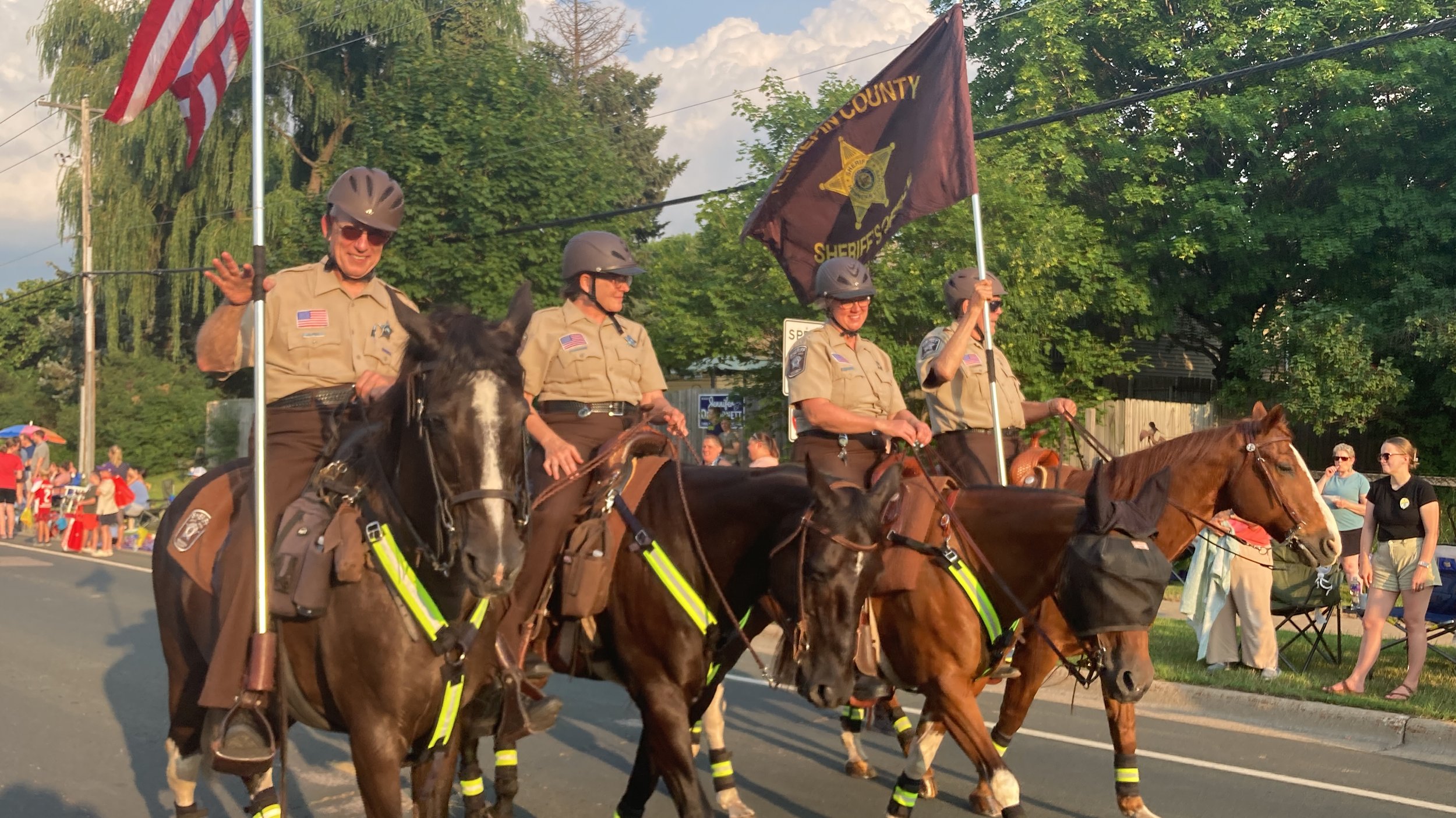 Four uniformed law enforcement officers riding horses in a parade. One officer is waving, and another is holding a flag that says 'Sheriff's Office.' There are spectators along the street, some with umbrellas, and green trees in the background.