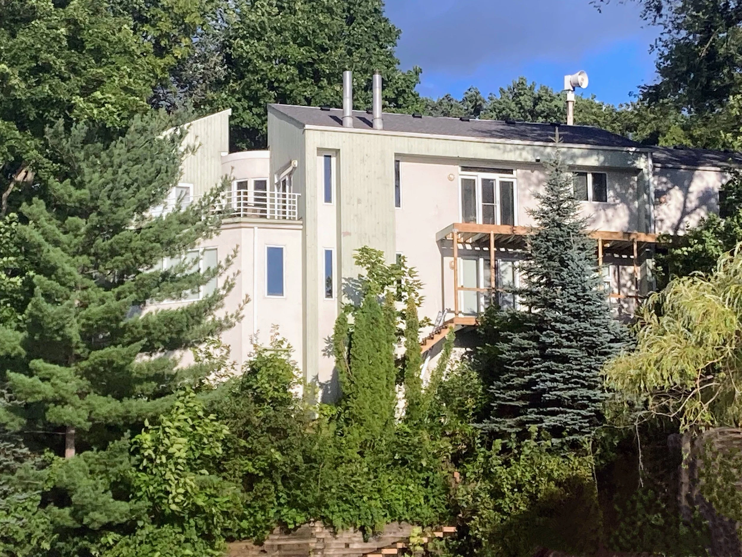A multi-story modern house surrounded by lush green trees, with a small wooden deck and large windows, under a partly cloudy blue sky.