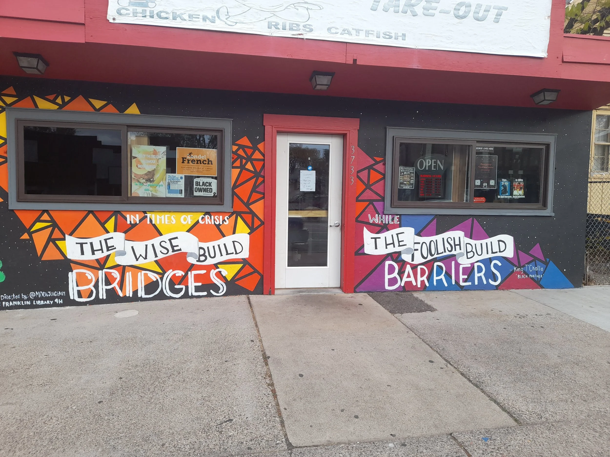 Colorful mural on building exterior with quotes about building bridges and barriers, featuring geometric patterns, windows, and a red door.
