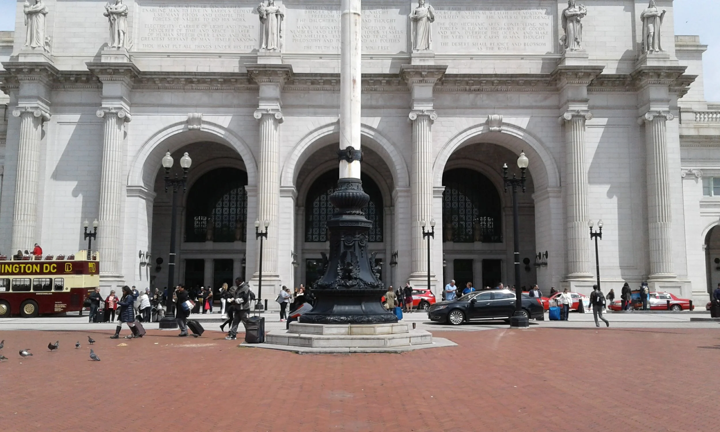 The entrance to a large, white, neoclassical building with tall columns and statues on top. There is a black street lamp in the center and people walking with luggage in front, along with several cars and a red and yellow double-decker bus on the sid