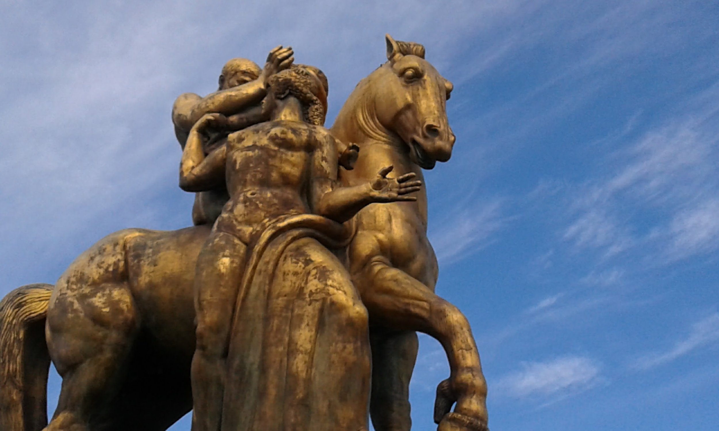 Bronze statue of a man on a horse, with two children riding on his back, set against a blue sky with wispy clouds.