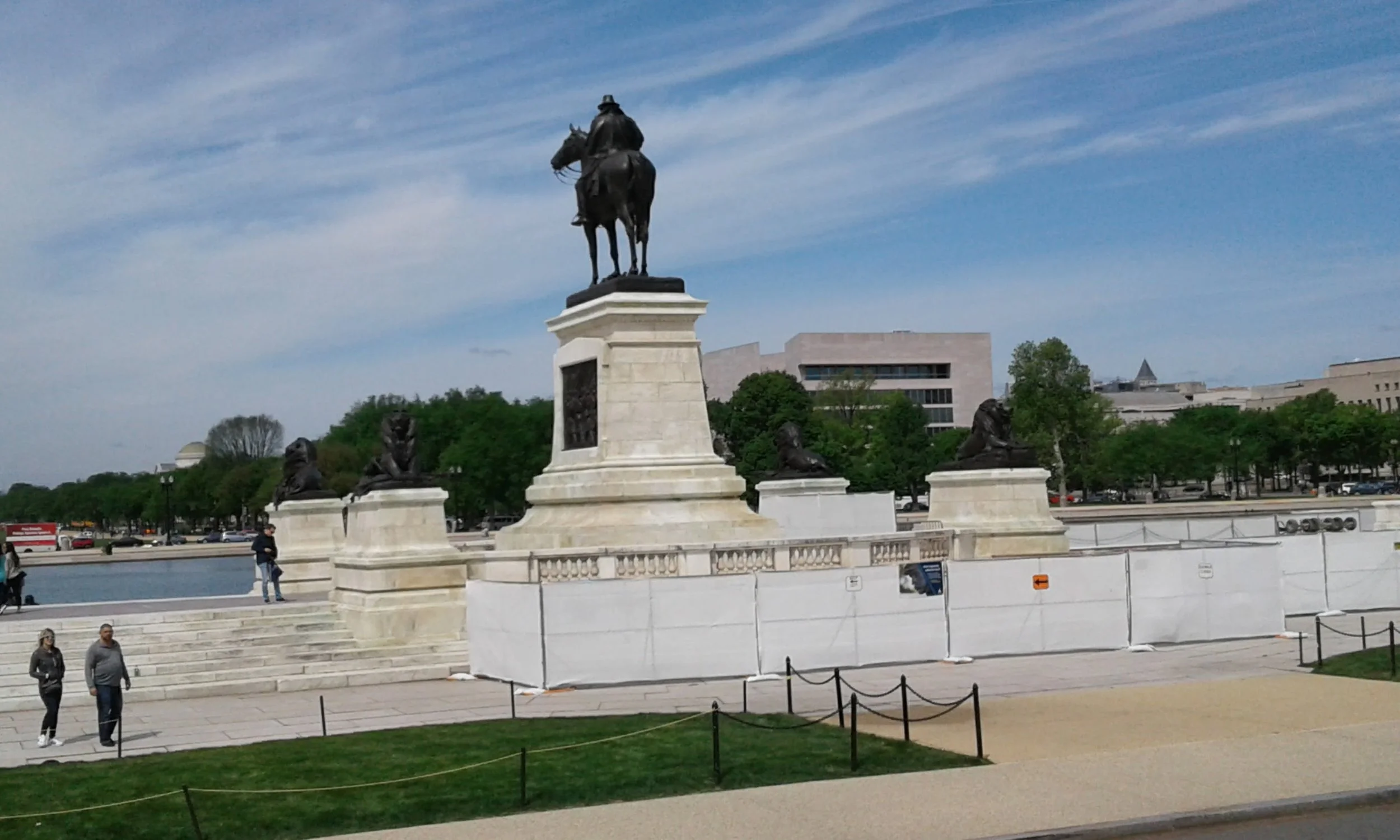 Statues of a man on a horse and four lions on pedestals in a park, with trees and buildings in the background under a partly cloudy sky.