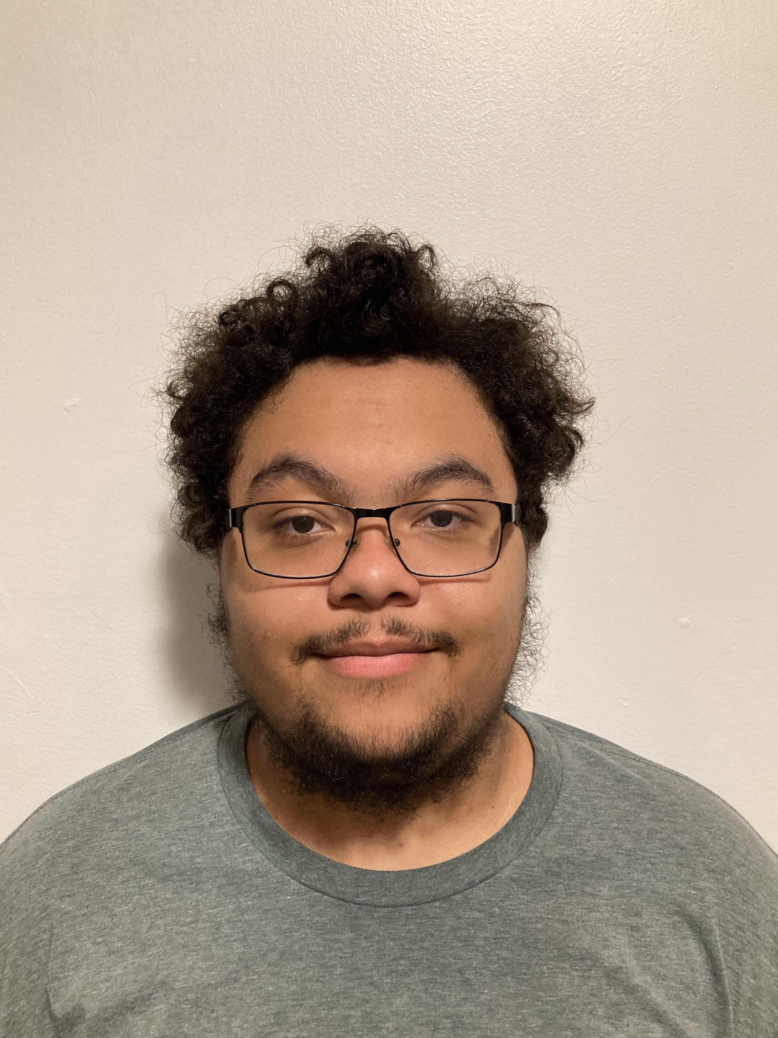 A young man with curly hair, glasses, and facial hair smiling at the camera against a plain white wall.
