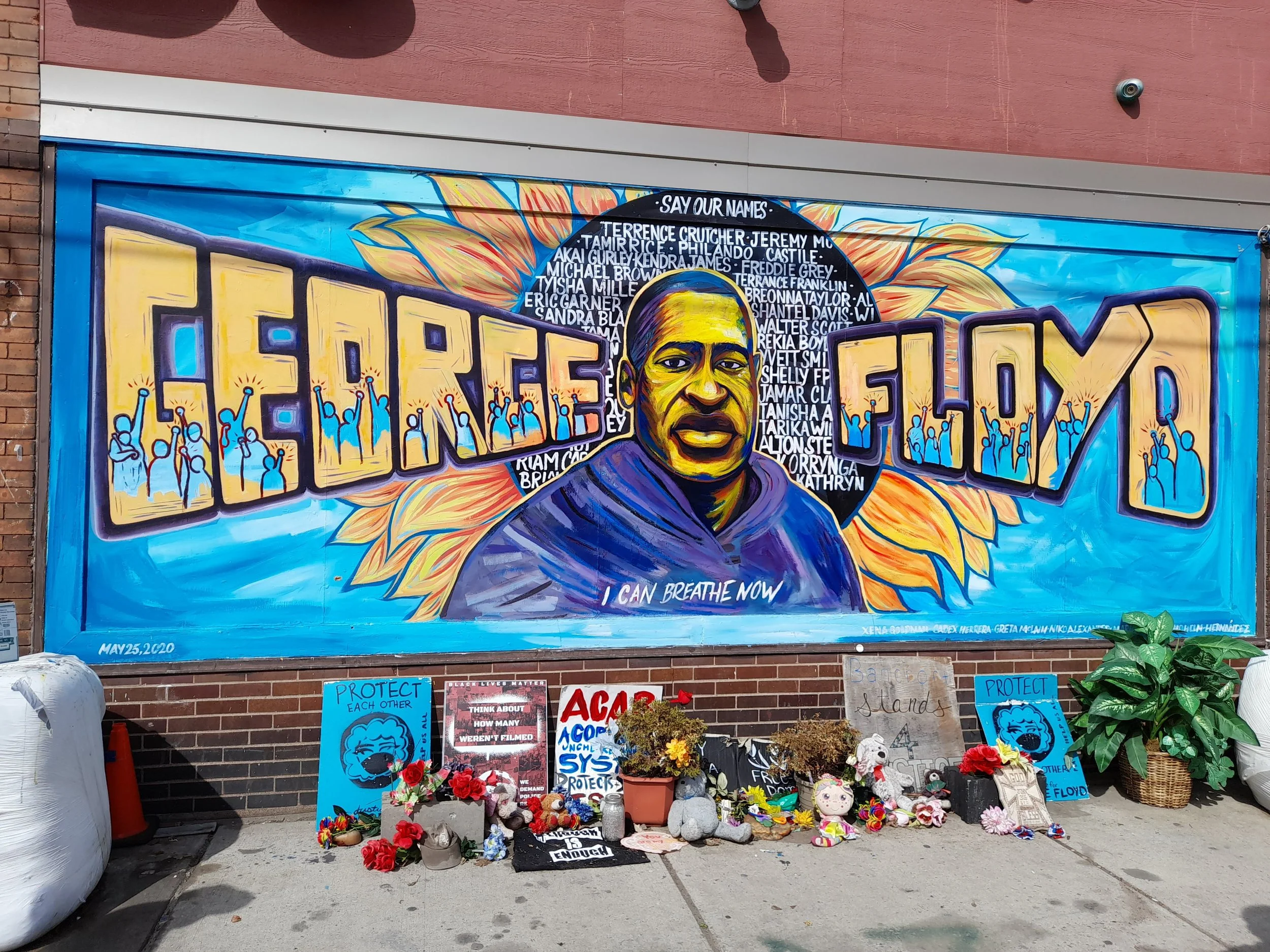 A mural honoring George Floyd featuring his portrait and the words 'GEORGE FLOYD' in large yellow letters, surrounded by an illustration of sunflowers and a background of blue sky. Beneath the portrait, the phrase 'I CAN BREATHE NOW' is written. In f