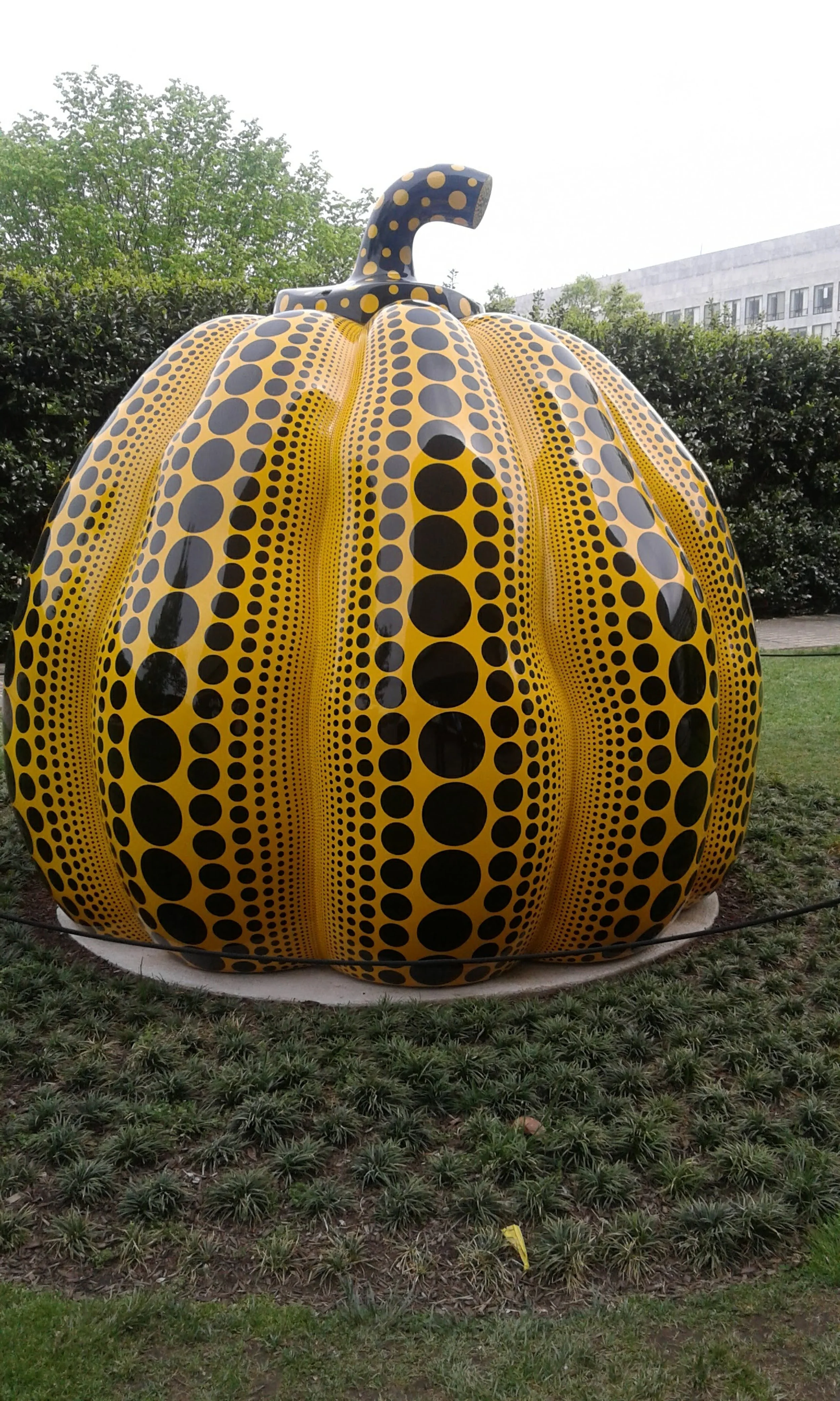 Large outdoor pumpkin sculpture with yellow and black dotted pattern, set on a grassy area with trees and a building in the background.