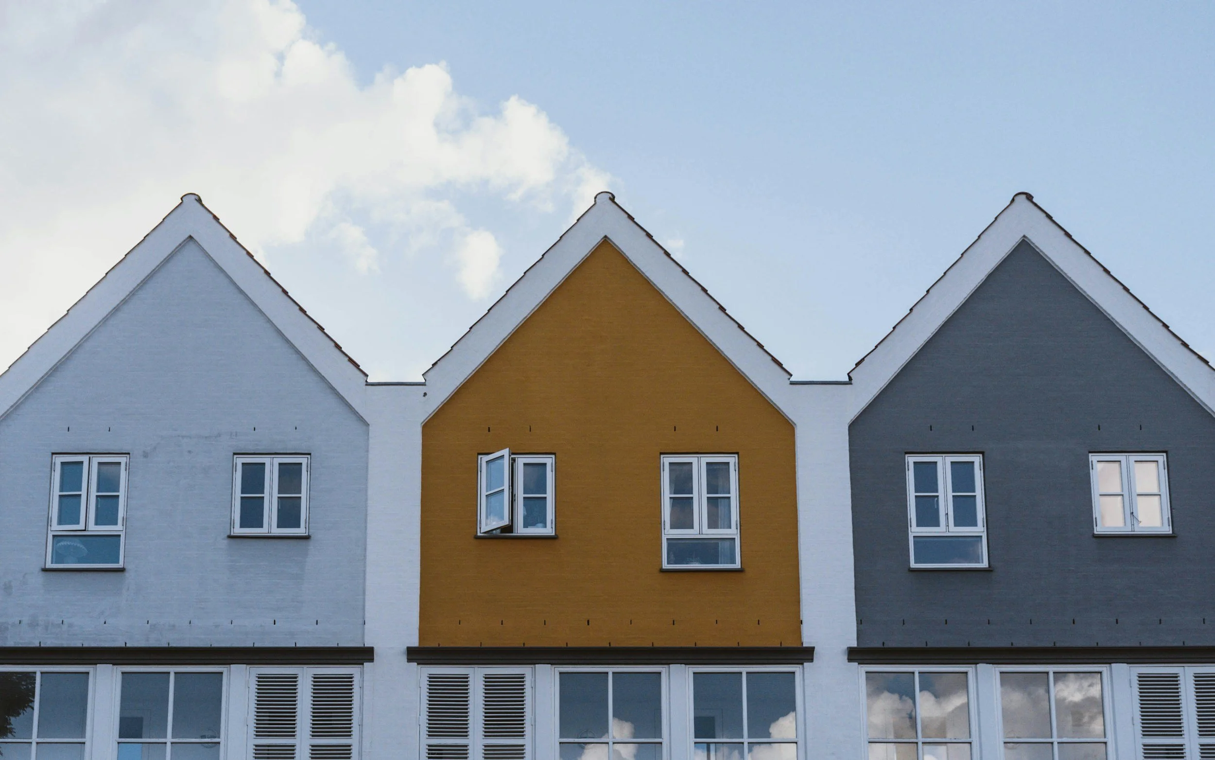 Colorful row of residential buildings with steep gabled roofs, painted in blue, yellow, and gray, with white window frames against a background of blue sky and some clouds.