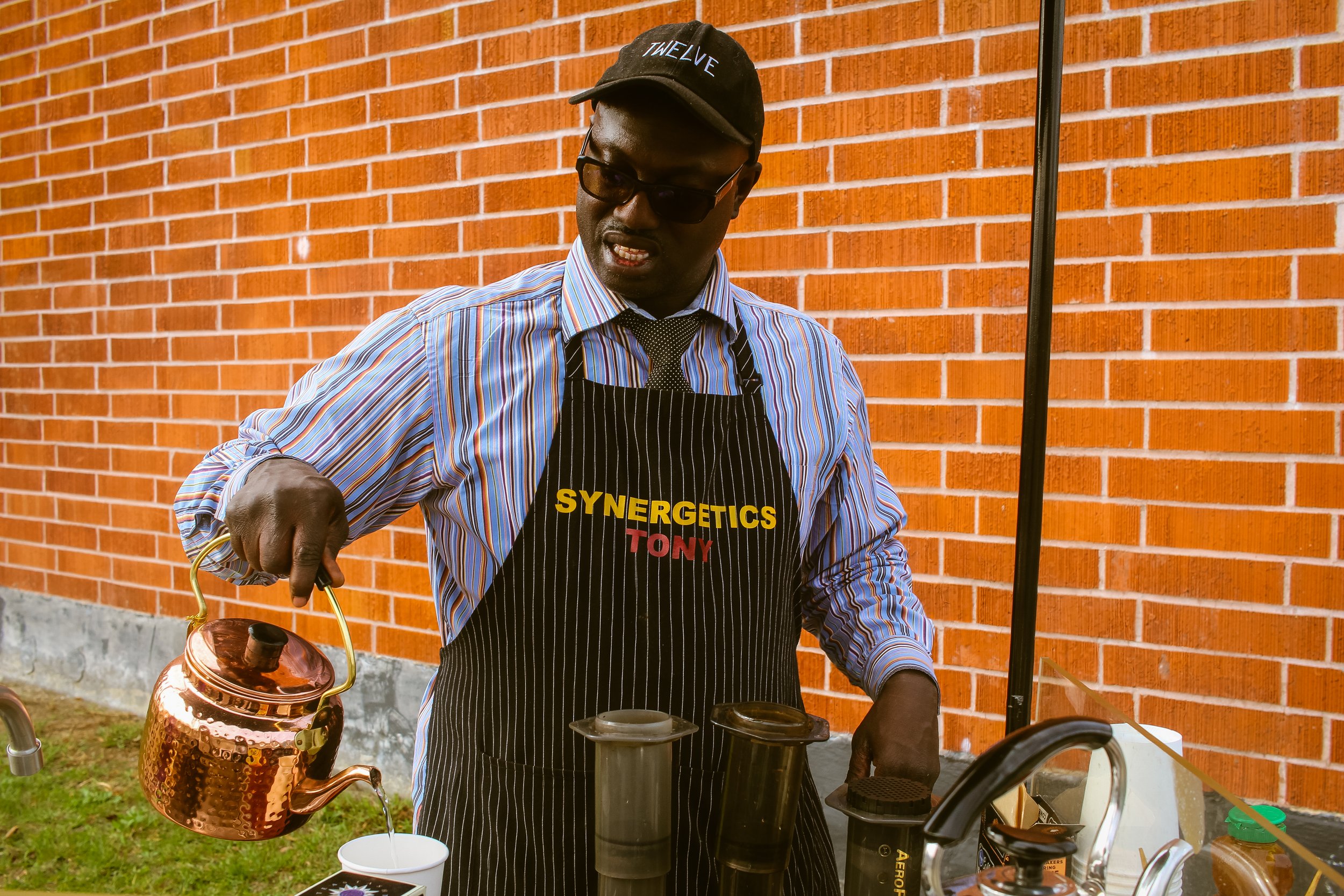Man pouring hot water from a copper kettle into a cup outdoors against a red brick wall. He is wearing a striped shirt, black apron with yellow and red lettering, black gloves, sunglasses, and a cap.