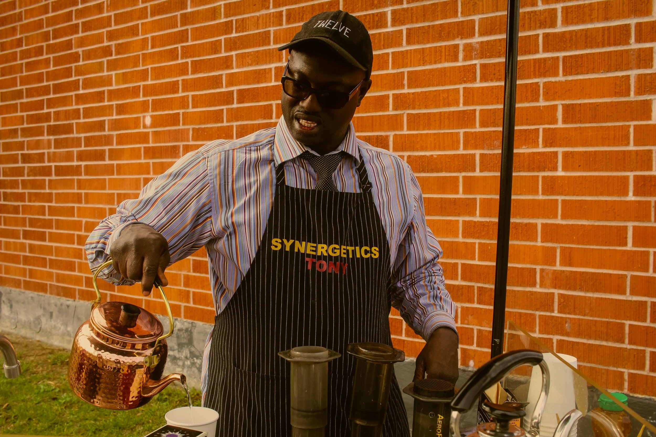A man in glasses wearing a striped shirt, apron, and a black cap pouring water from a copper kettle into a cup.