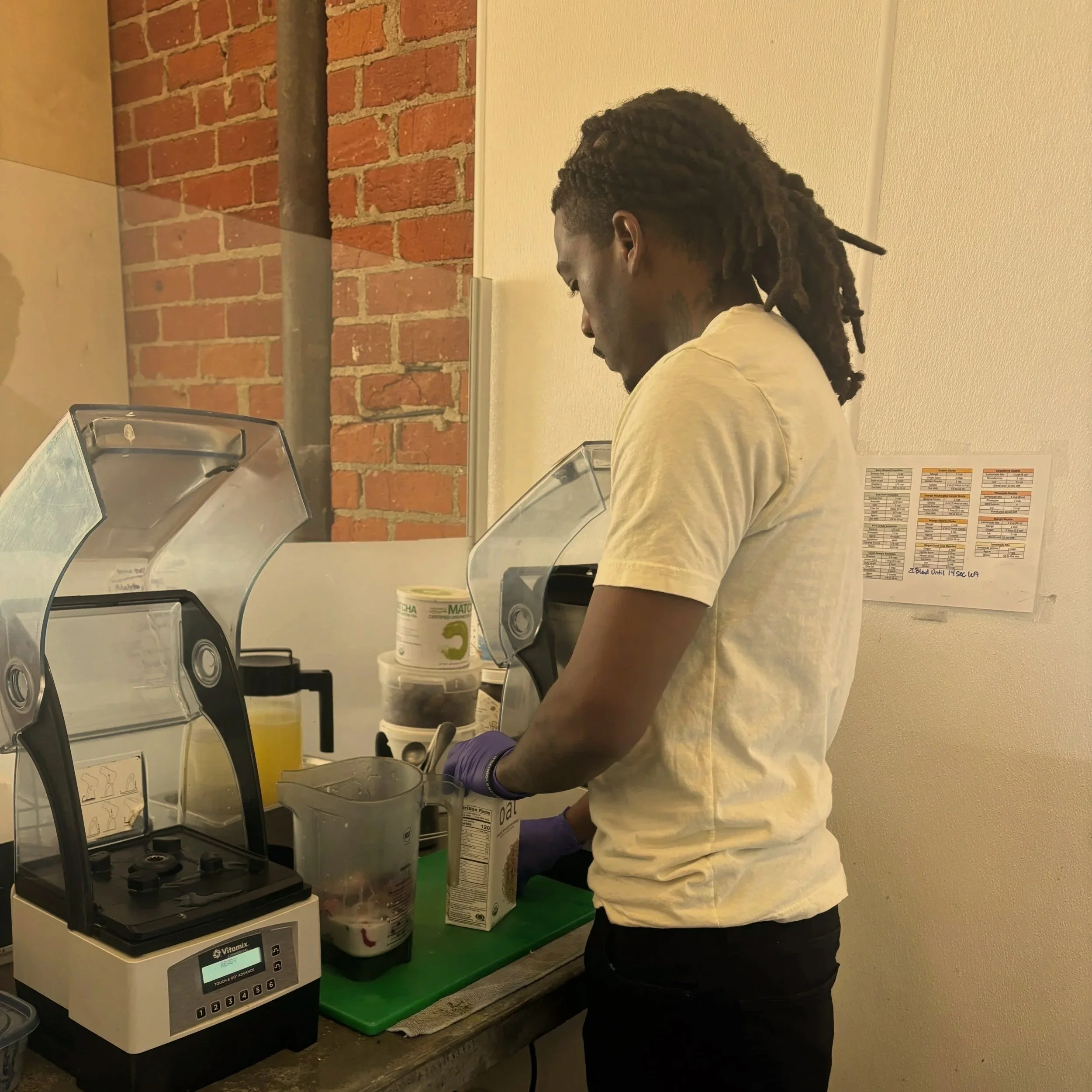 Man with dreadlocks wearing a beige t-shirt and purple gloves preparing ingredients in a kitchen with a blender and containers.