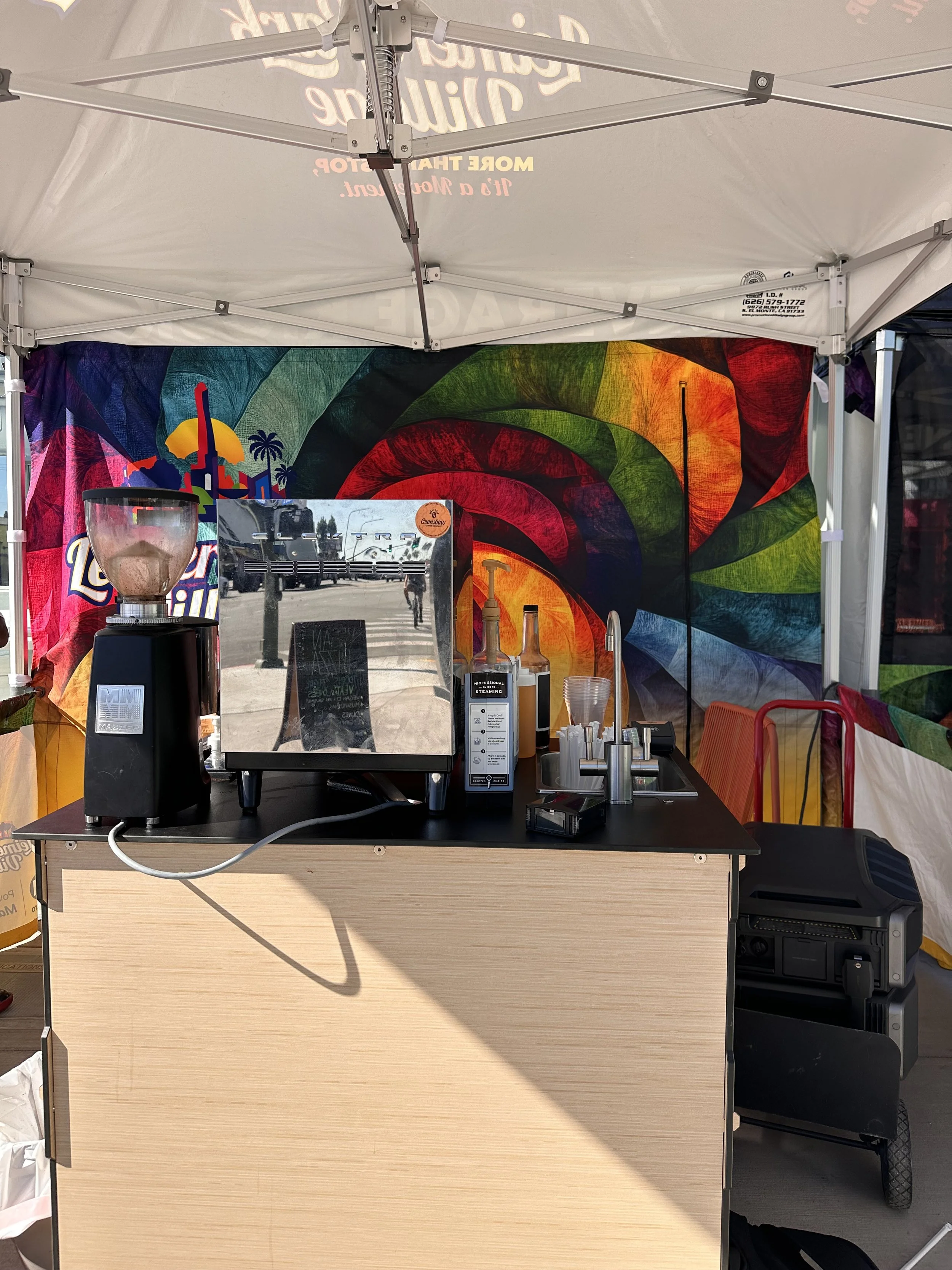 A colorful outdoor food or drink stand with a black countertop, a blender, and various bottles and cups. Behind the stand, there is a vibrant, spiraling rainbow-colored backdrop under a white canopy.