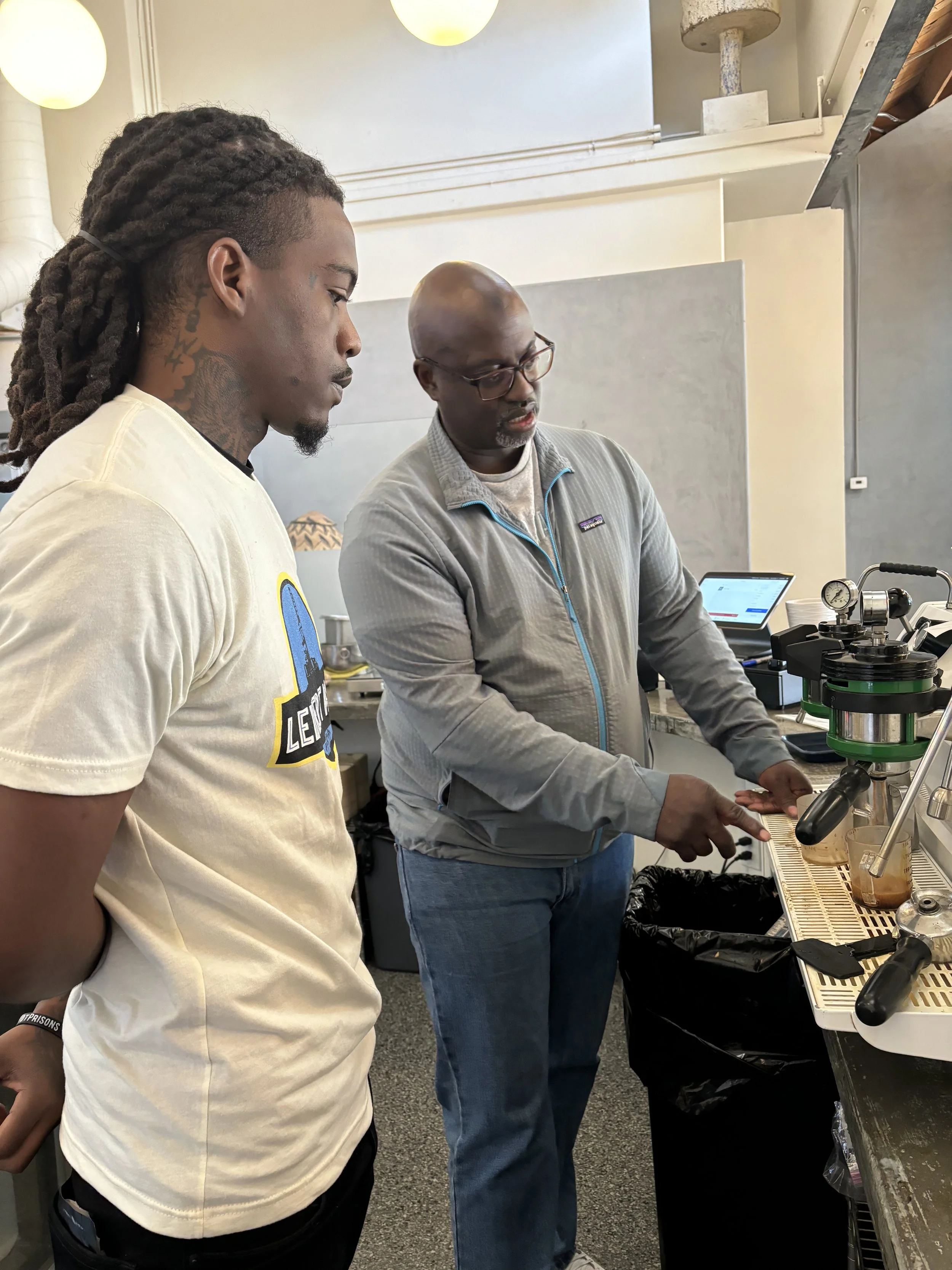 Two men are working with coffee brewing equipment in a kitchen or café setting. One man, wearing a grey Patagonia jacket and glasses, is explaining or demonstrating something about the coffee setup to the other man, who has long dreadlocks and a tattoo on his neck, wearing a white t-shirt with a logo.
