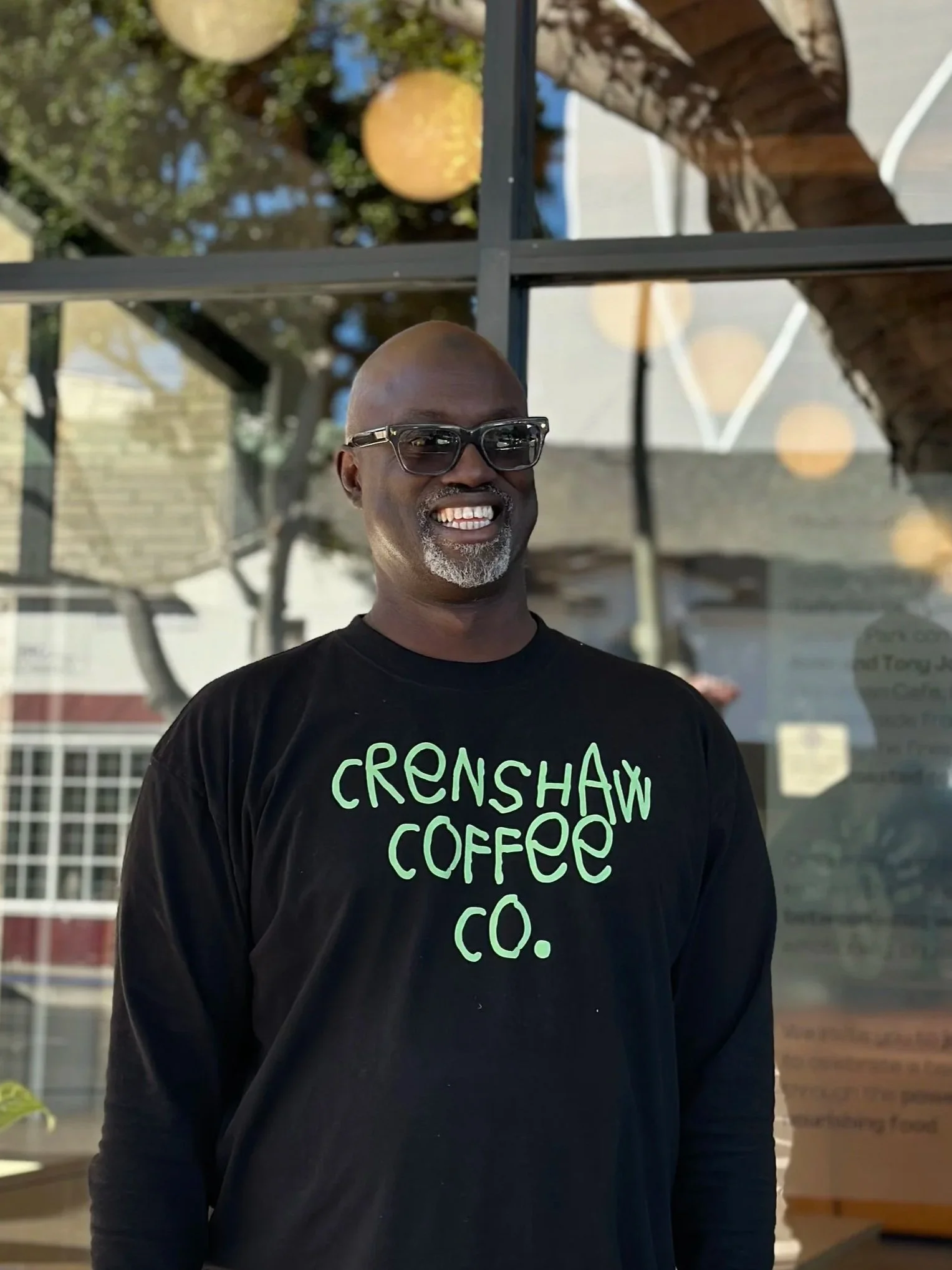 A smiling man wearing glasses and a black shirt with green text that reads 'Crenshaw Coffee Co.' standing outdoors in front of a window and tree.