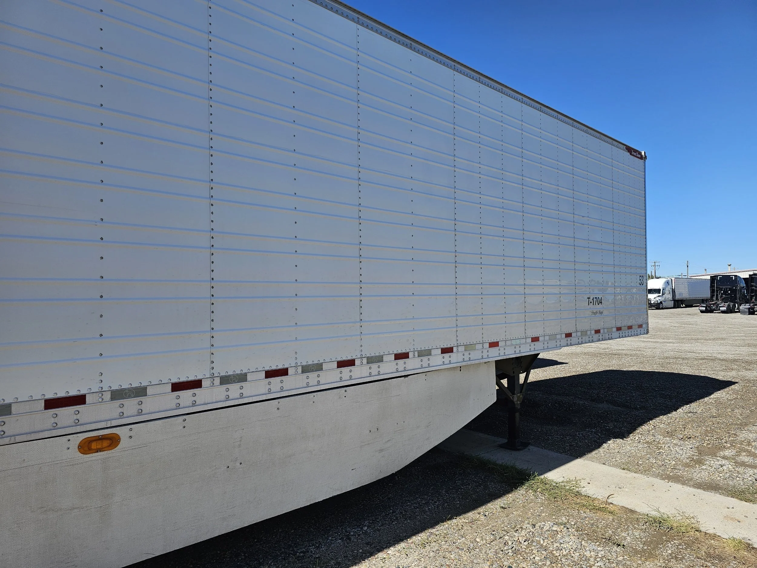 A large white semi-trailer truck parked on a gravel lot under a clear blue sky, with other trucks visible in the background.