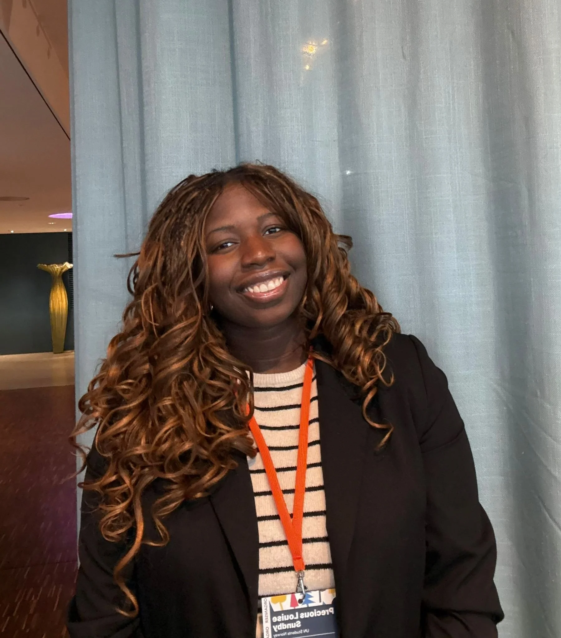 A young woman with long, curly, brown hair smiling at the camera, wearing a black blazer over a striped top, with a conference badge hanging around her neck, standing in front of a light-colored curtain.
