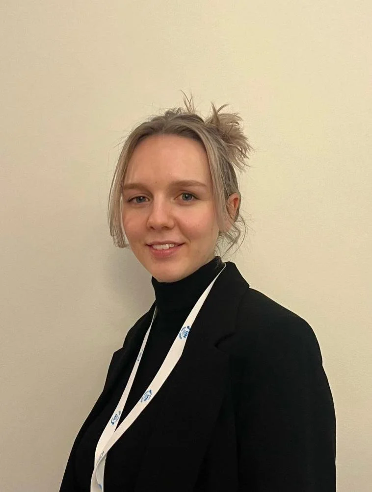 A young woman with blonde hair in a messy bun, wearing a black blazer and turtleneck, smiling in front of a plain beige wall, with a white lanyard around her neck.