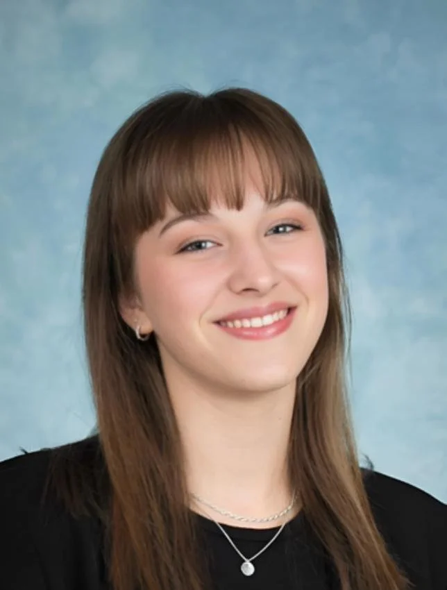A young woman with straight brown hair, bangs, and light makeup smiling in front of a blue background.