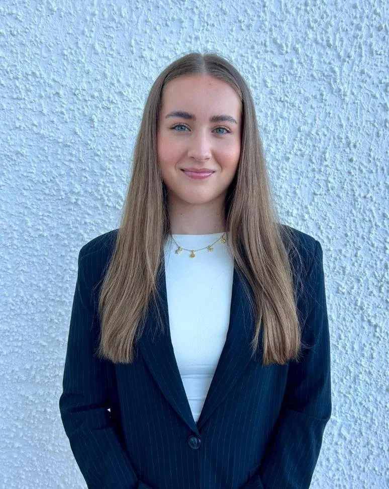 A young woman with long, straight brown hair and blue eyes, wearing a dark pinstripe blazer over a white top and a gold necklace, standing against a textured white wall.