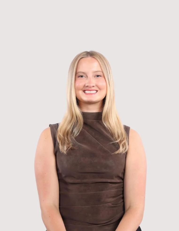 A young woman with long blonde hair, smiling, wearing a sleeveless brown top, standing against a plain light gray background.